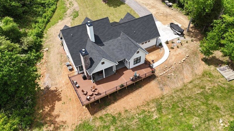 Aerial view of a white farmhouse with a dark roof and a large wooden deck. Surrounded by green trees and grass.