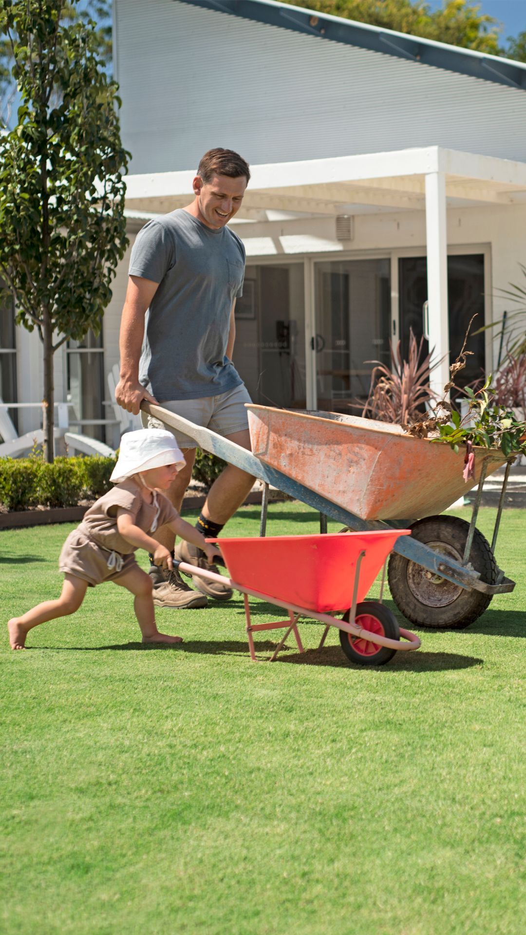 Man Pushing a Wheelbarrow Across a Lawn While a Child Runs Beside Him Near a House — Tweed Landscape Supplies in Tweed Heads, NSW