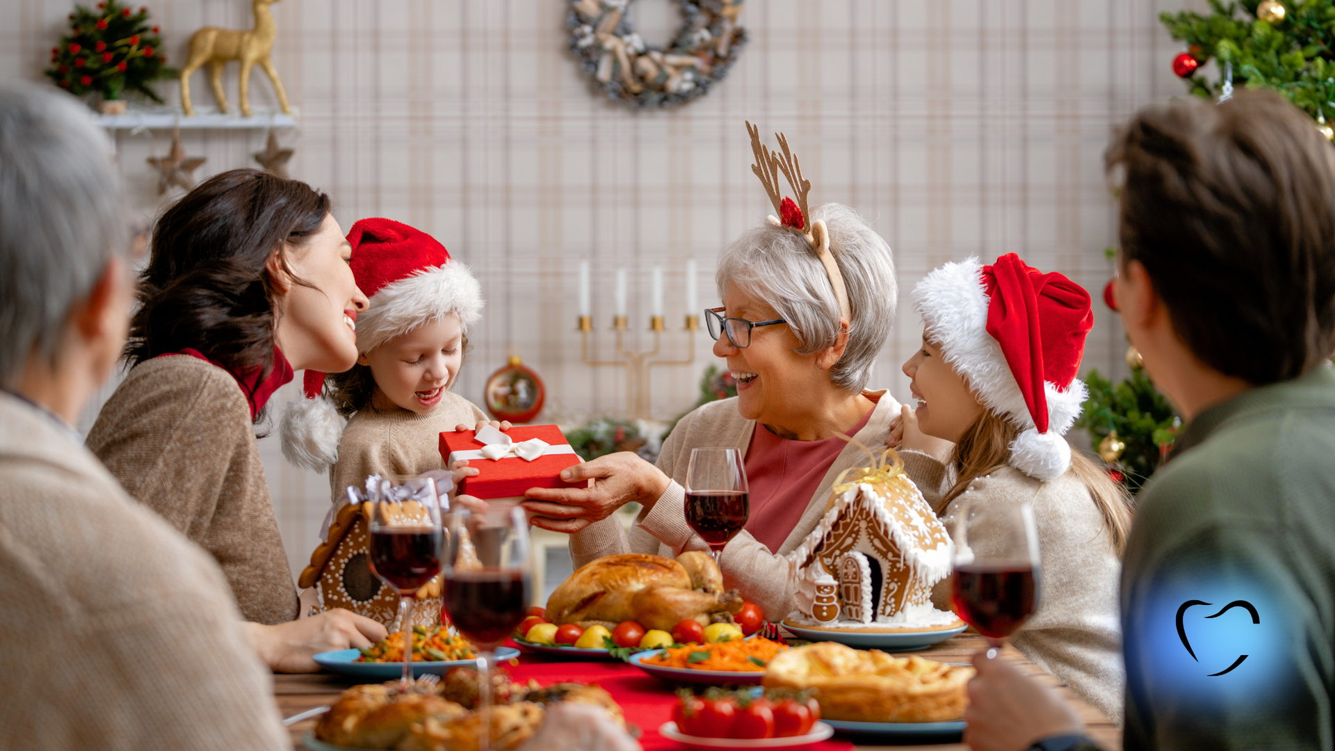 A family shares a festive holiday meal, with a child handing a small gift to an older family member at a table.