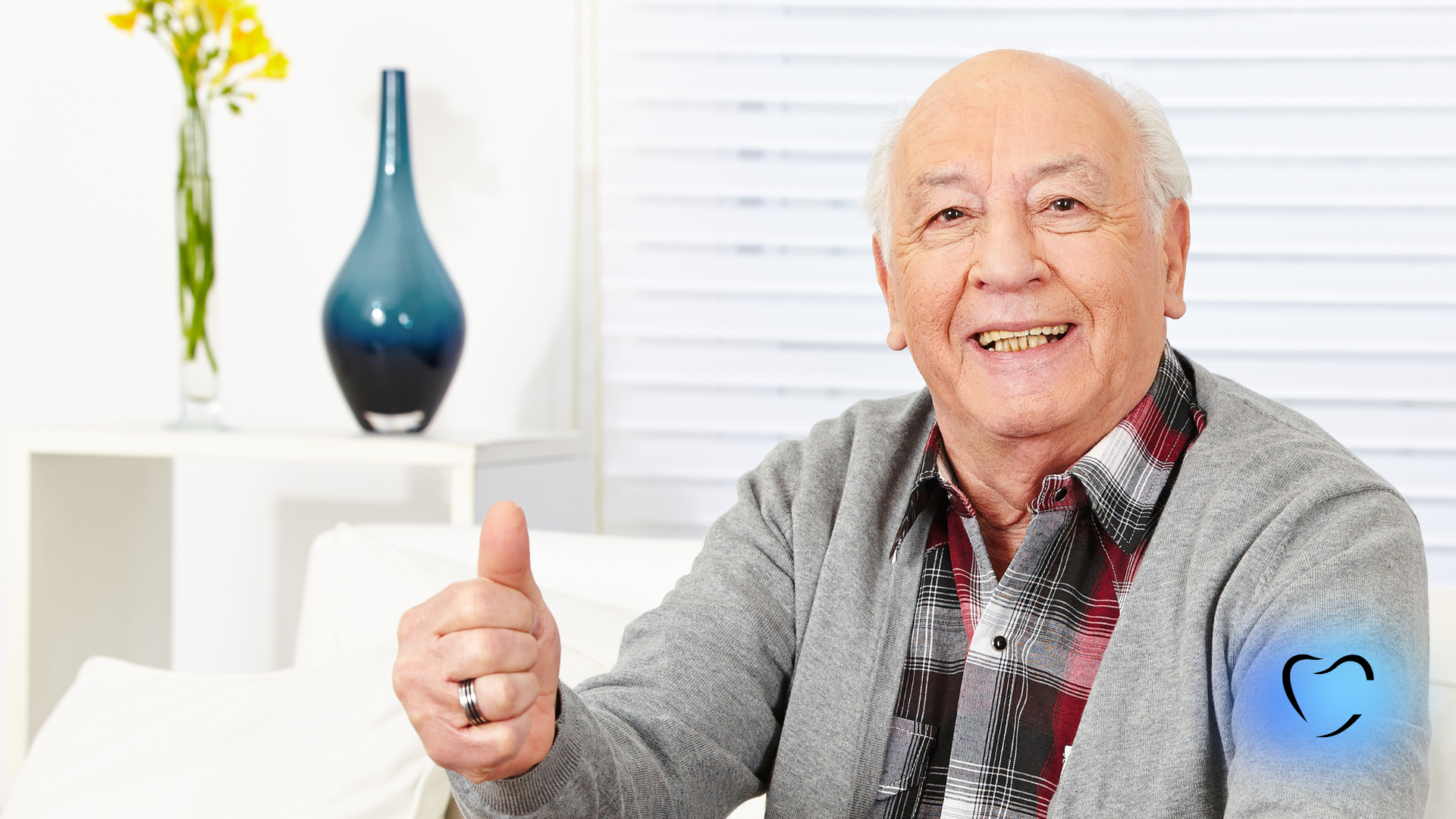 A person in a gray sweater and plaid shirt sits indoors, smiling and giving a thumbs up.