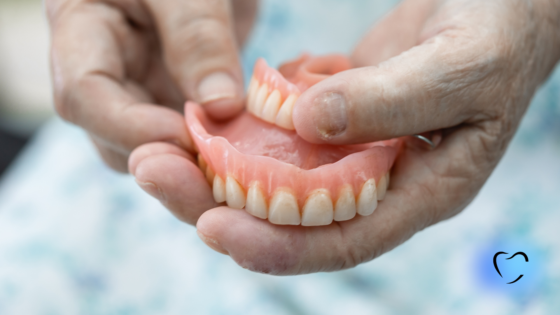 Close-up of hands holding upper and lower sets of dentures against a light blue patterned background.