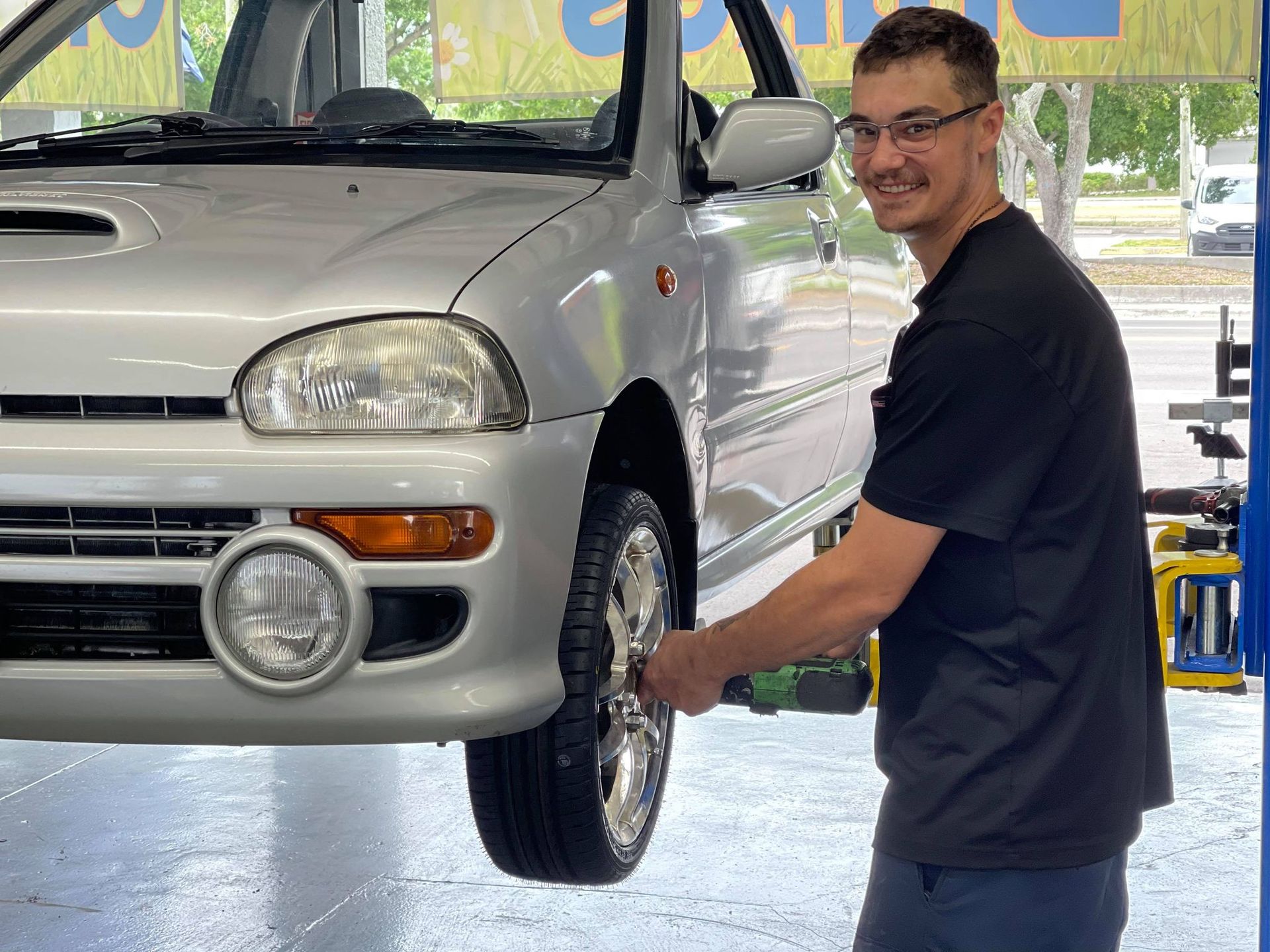 Man working on the front wheel of a white car in a garage, smiling at the camera. | Gulf Coast European Automotive