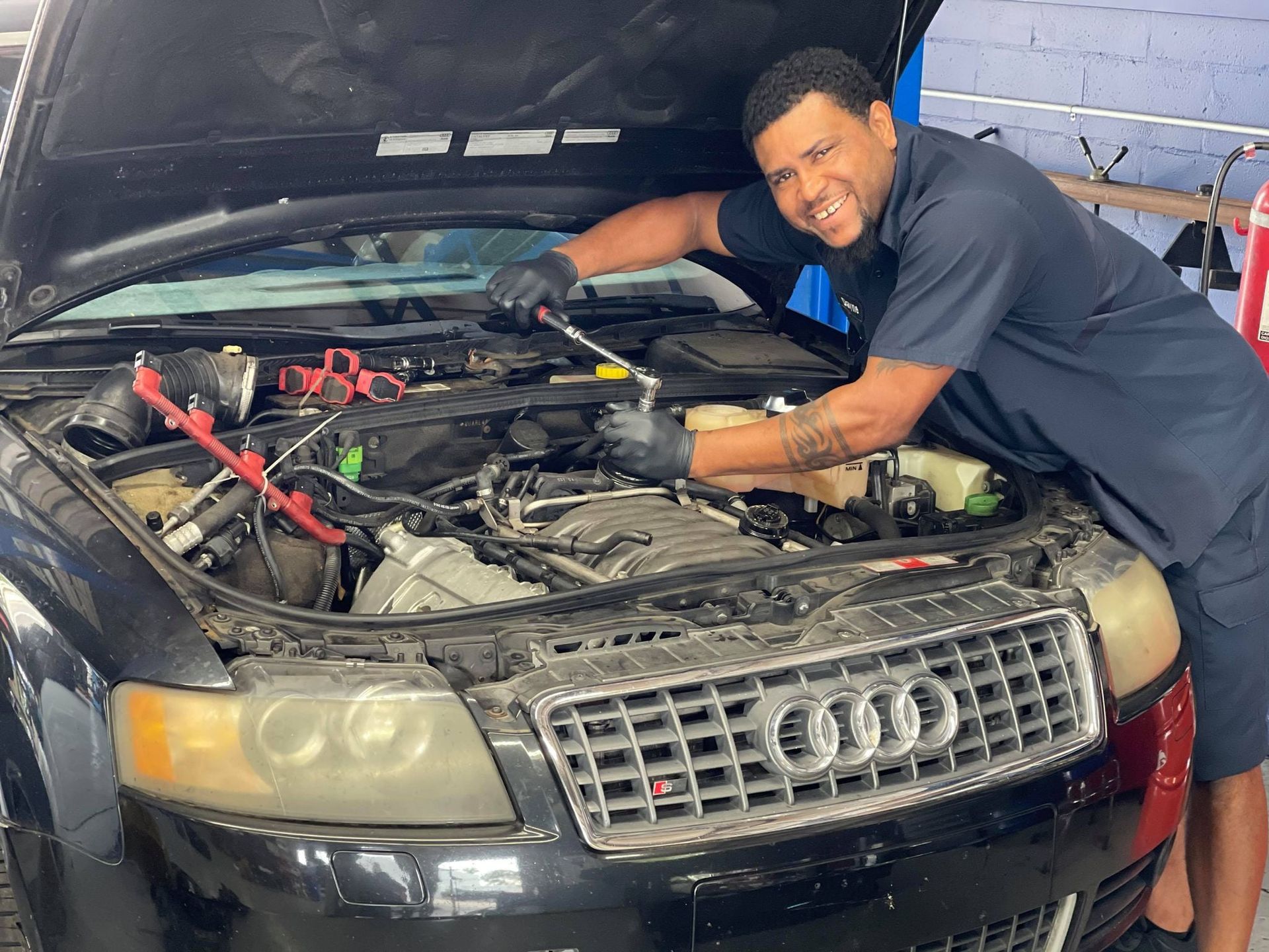 Mechanic working under the hood of a black Audi in a garage, smiling at the camera | Gulf Coast European Automotive