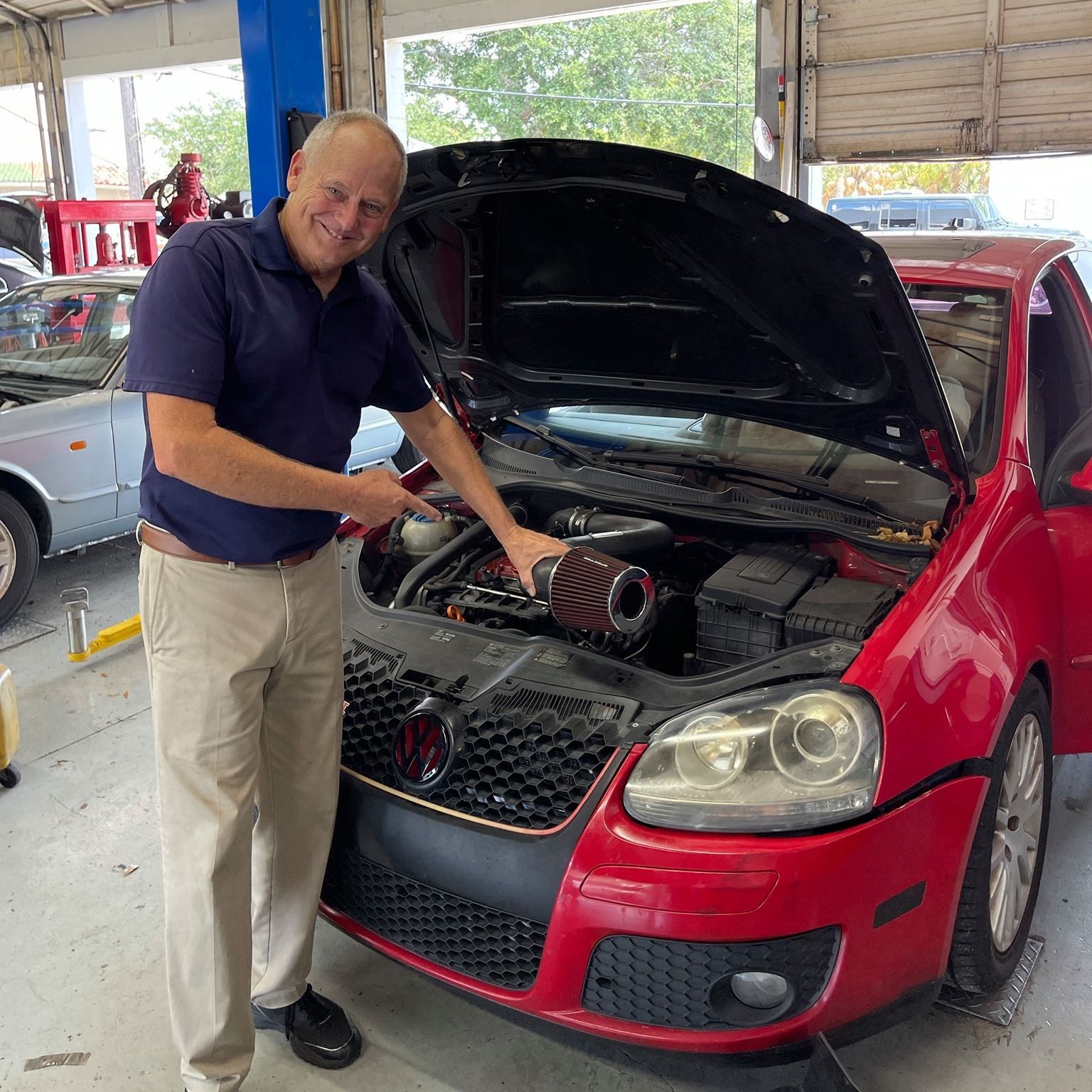 Man working on a red car with hood open in a garage, holding a tool near the engine. | Gulf Coast European Automotive