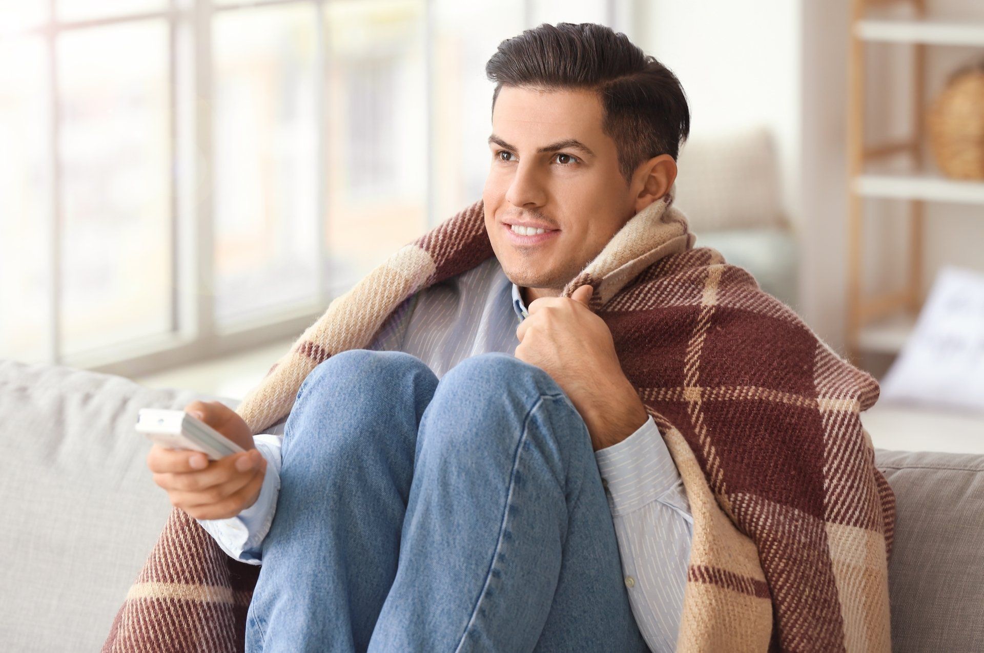 Un homme enveloppé dans une couverture est assis sur un canapé tenant une télécommande.
