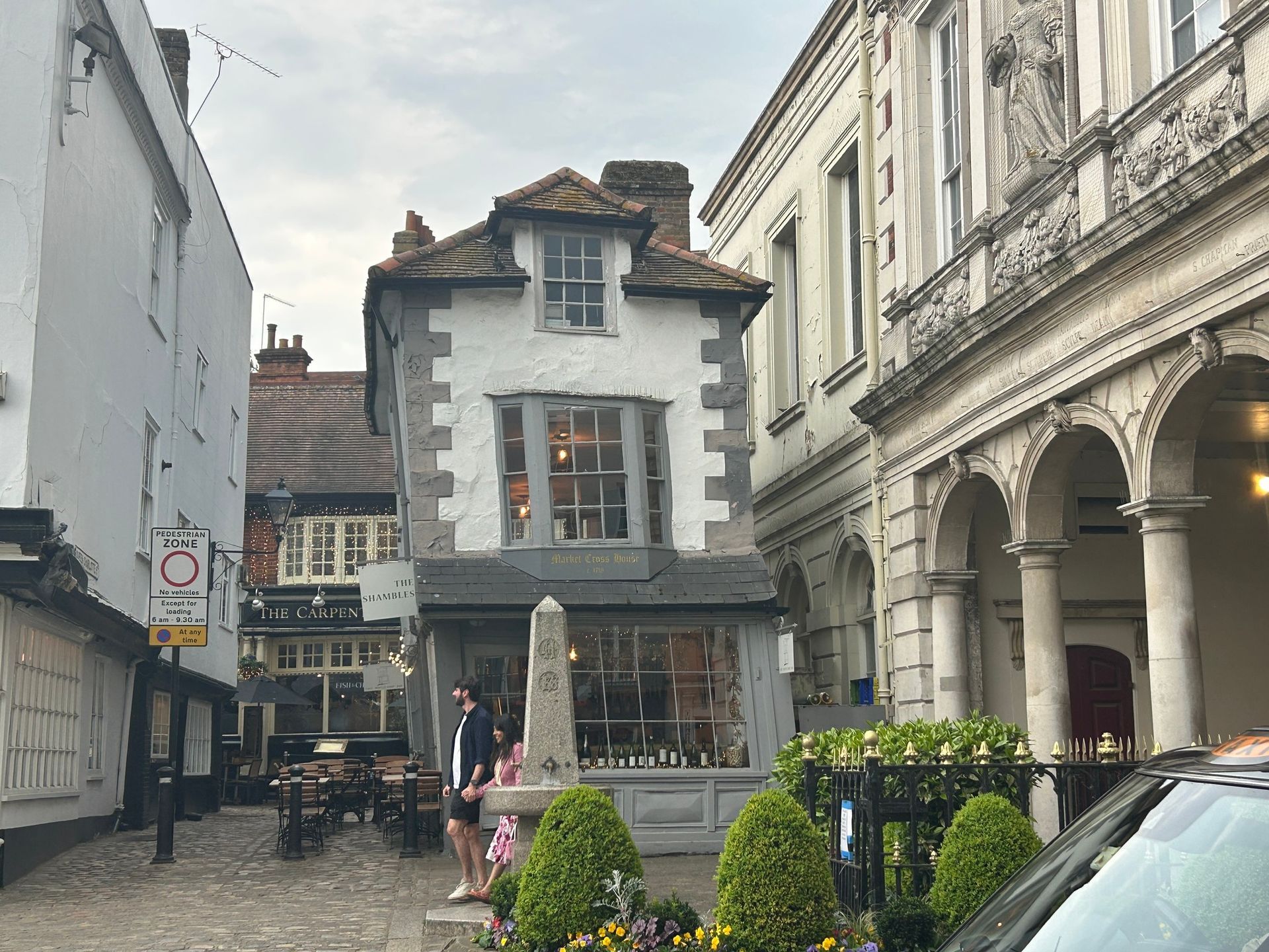 Narrow European street with old buildings, café seating, and a stone monument in the center