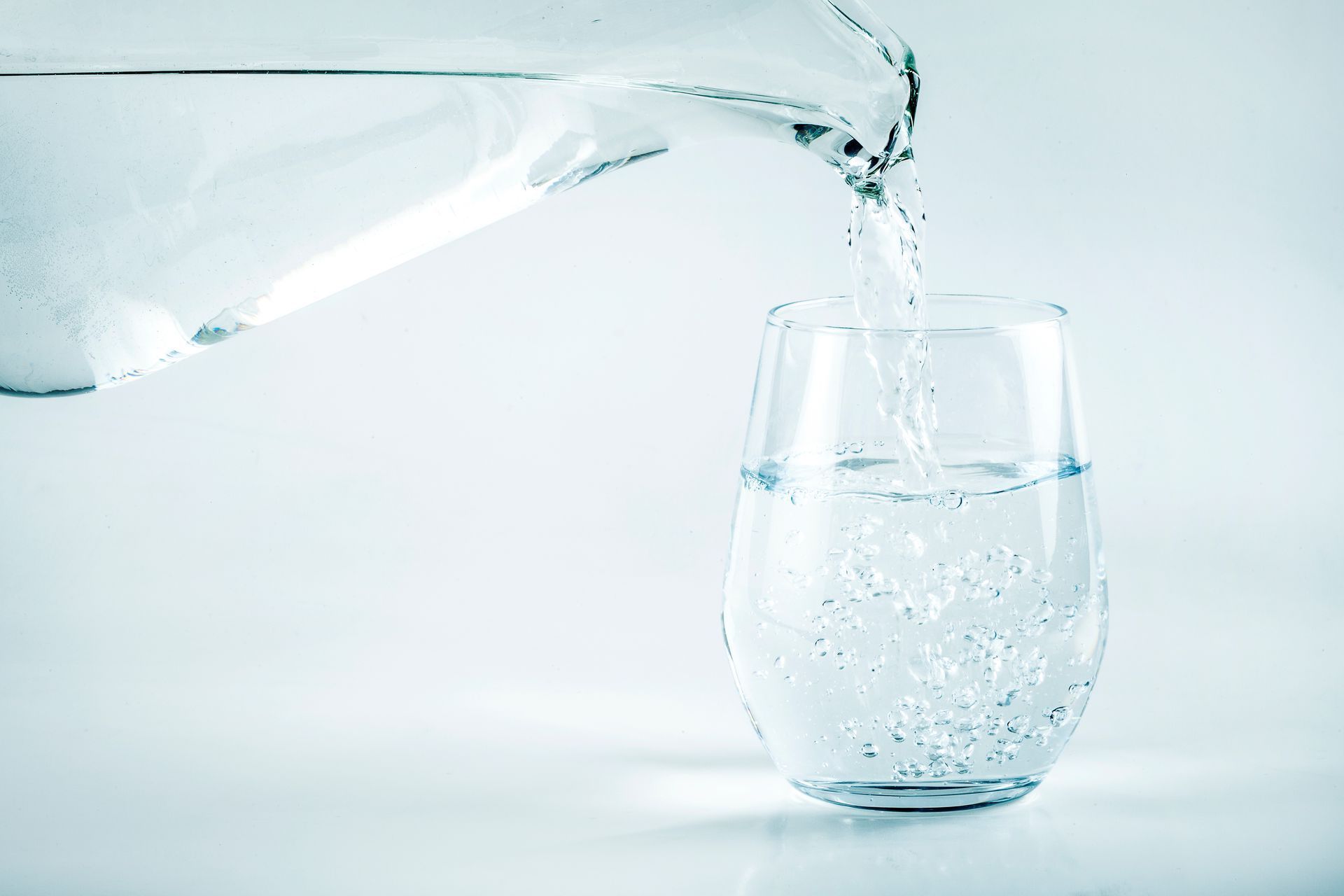 Water being poured from a pitcher into a glass, both are clear, against a white background.