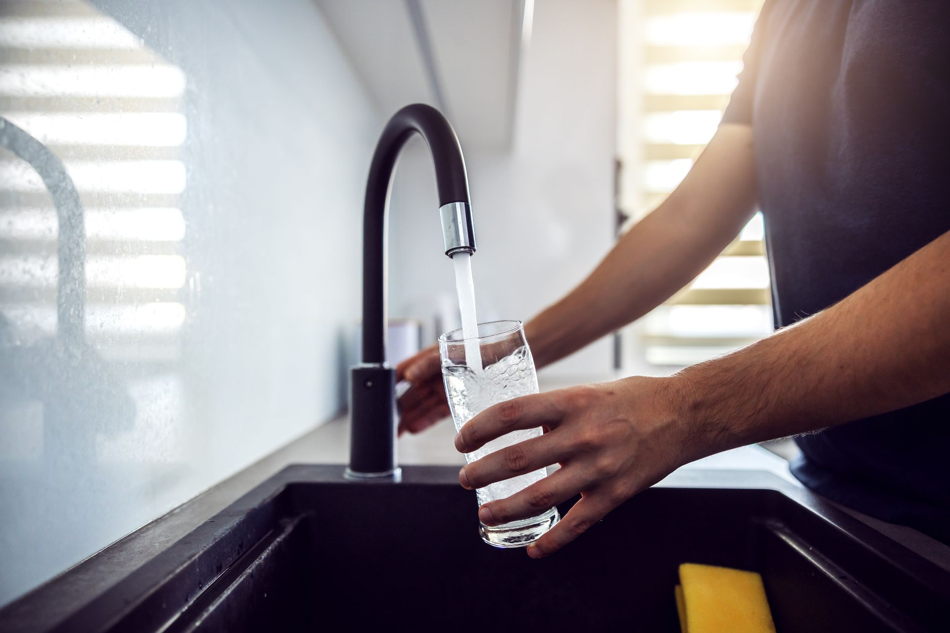 Person filling a glass with water from a kitchen faucet. Black sink, blue shirt.