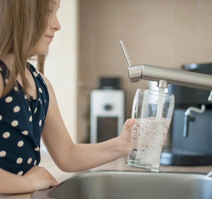 Girl filling a glass with water from a kitchen faucet over a sink.