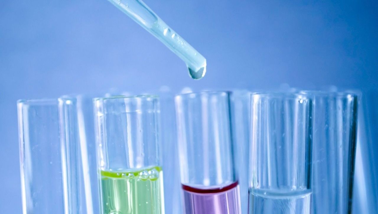 Dropper dispensing liquid into test tubes, some containing colored solutions, in a lab setting.