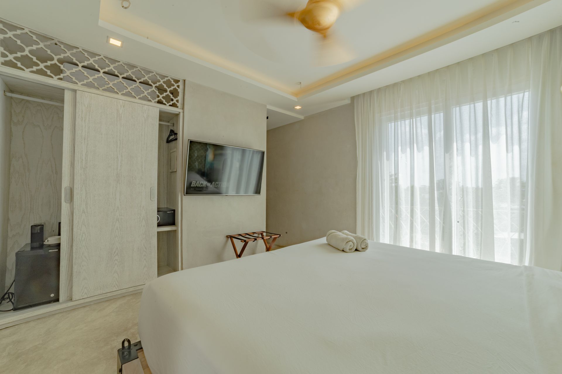Bedroom with white bed, closet, TV, sheer curtains, ceiling fan.