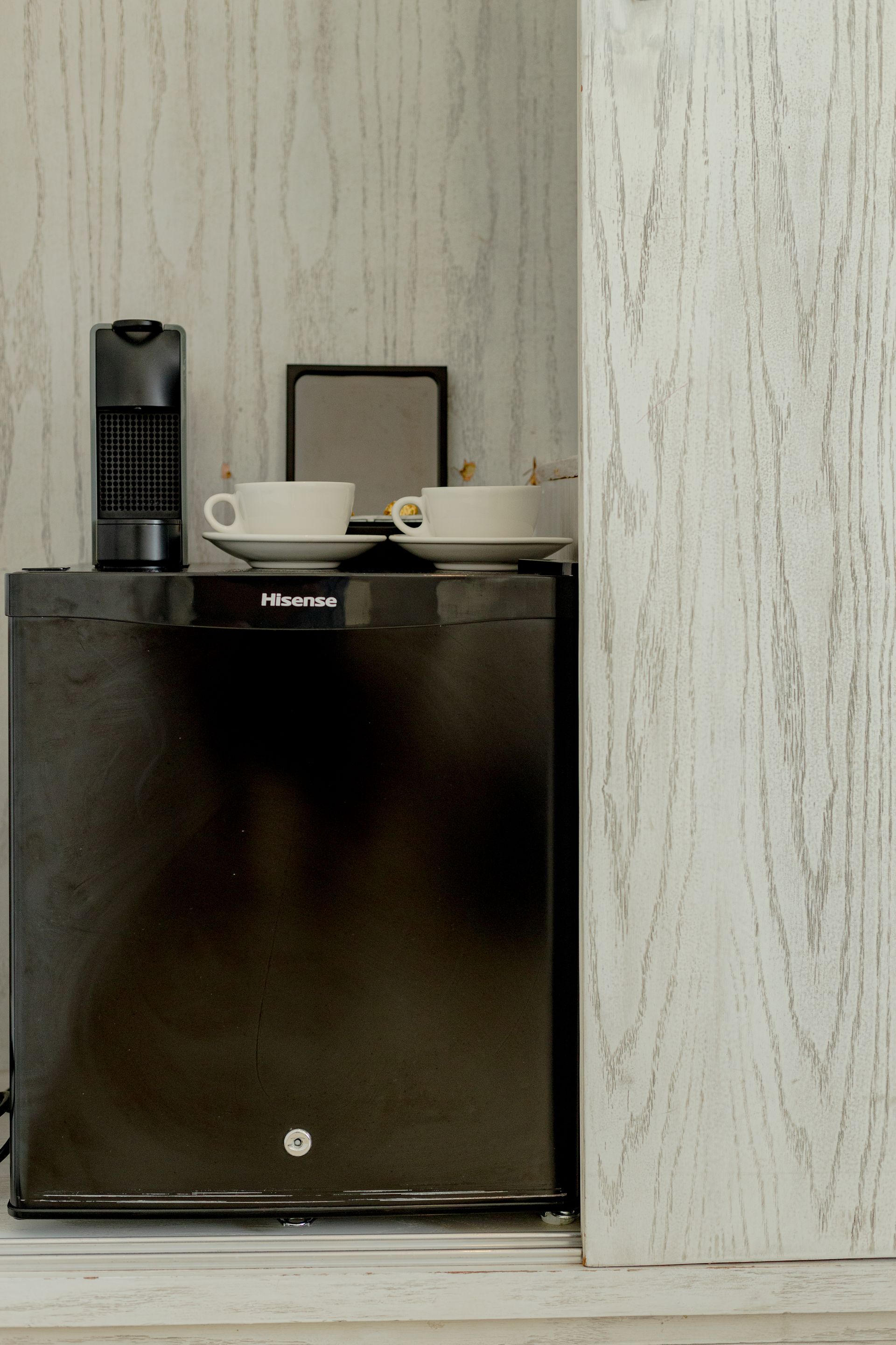 Black mini-fridge with coffee cups, a coffee maker, and a tray on top, against a light wood-paneled wall.