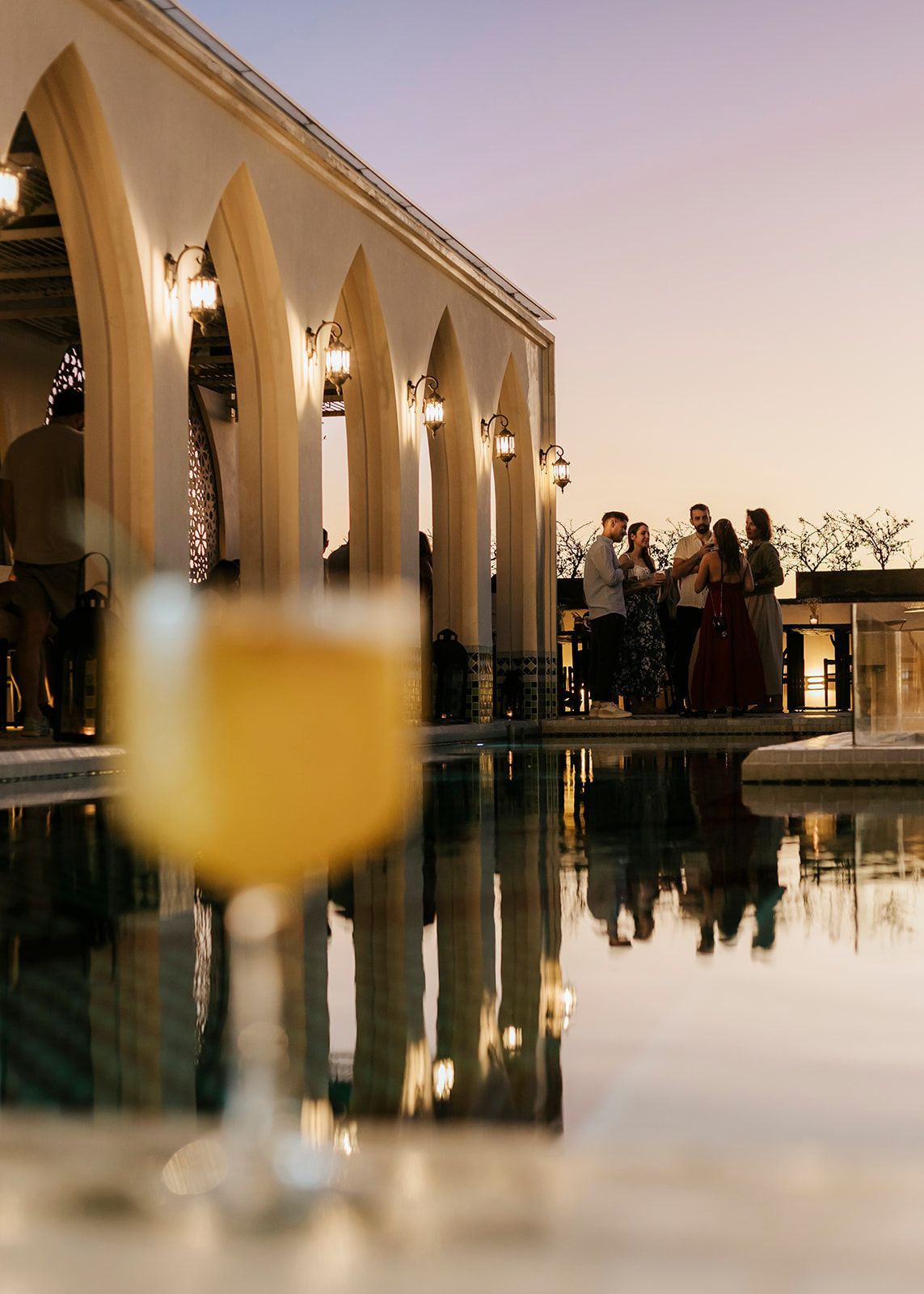 A group of people are standing around a pool with a glass of wine in the foreground.