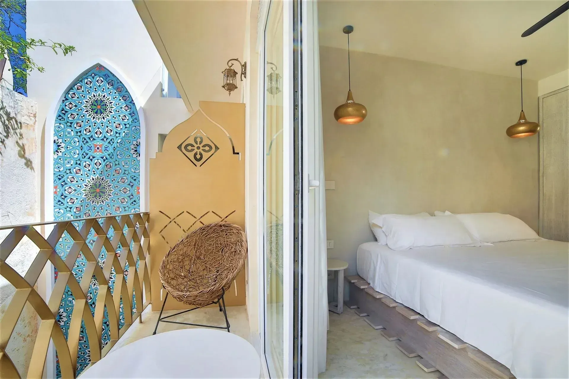 Bedroom with balcony view: beige walls, white bed, gold light fixtures, rattan chair, turquoise mosaic.