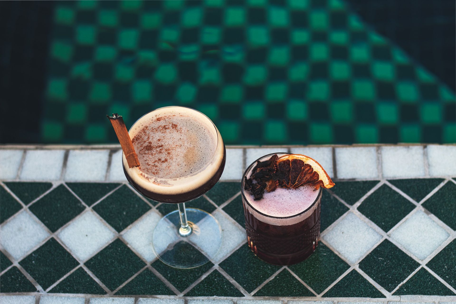Two drinks are sitting on a tiled table next to a pool.