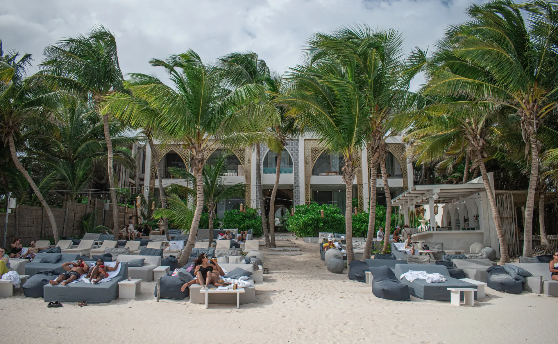 Beachside lounge with palm trees, a white building, and people relaxing on cushioned seating.