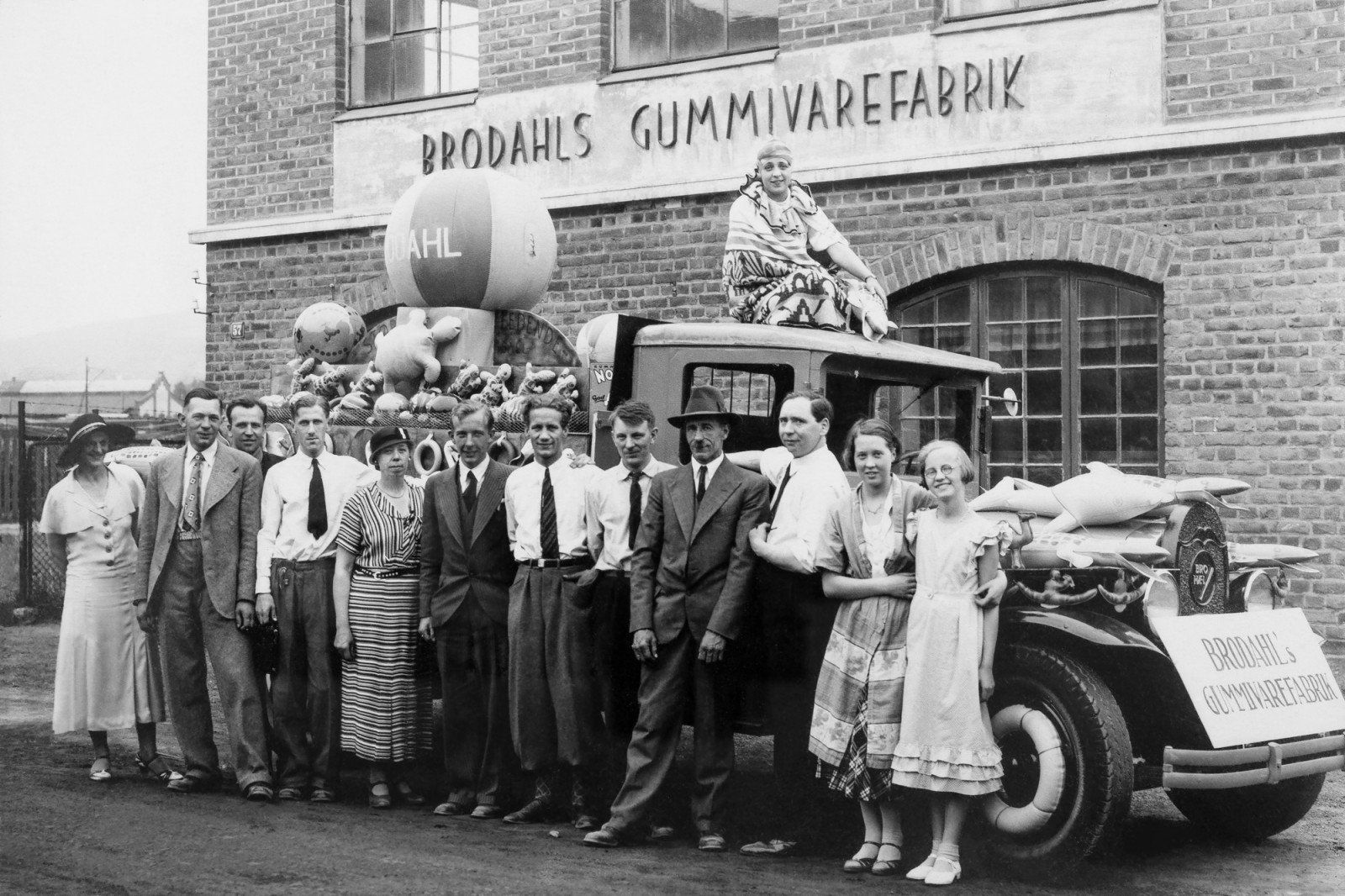 Gruppe mennesker poserer med bil foran bygningen «Brodahls Gummivarefabrik», ballonger på bilen.