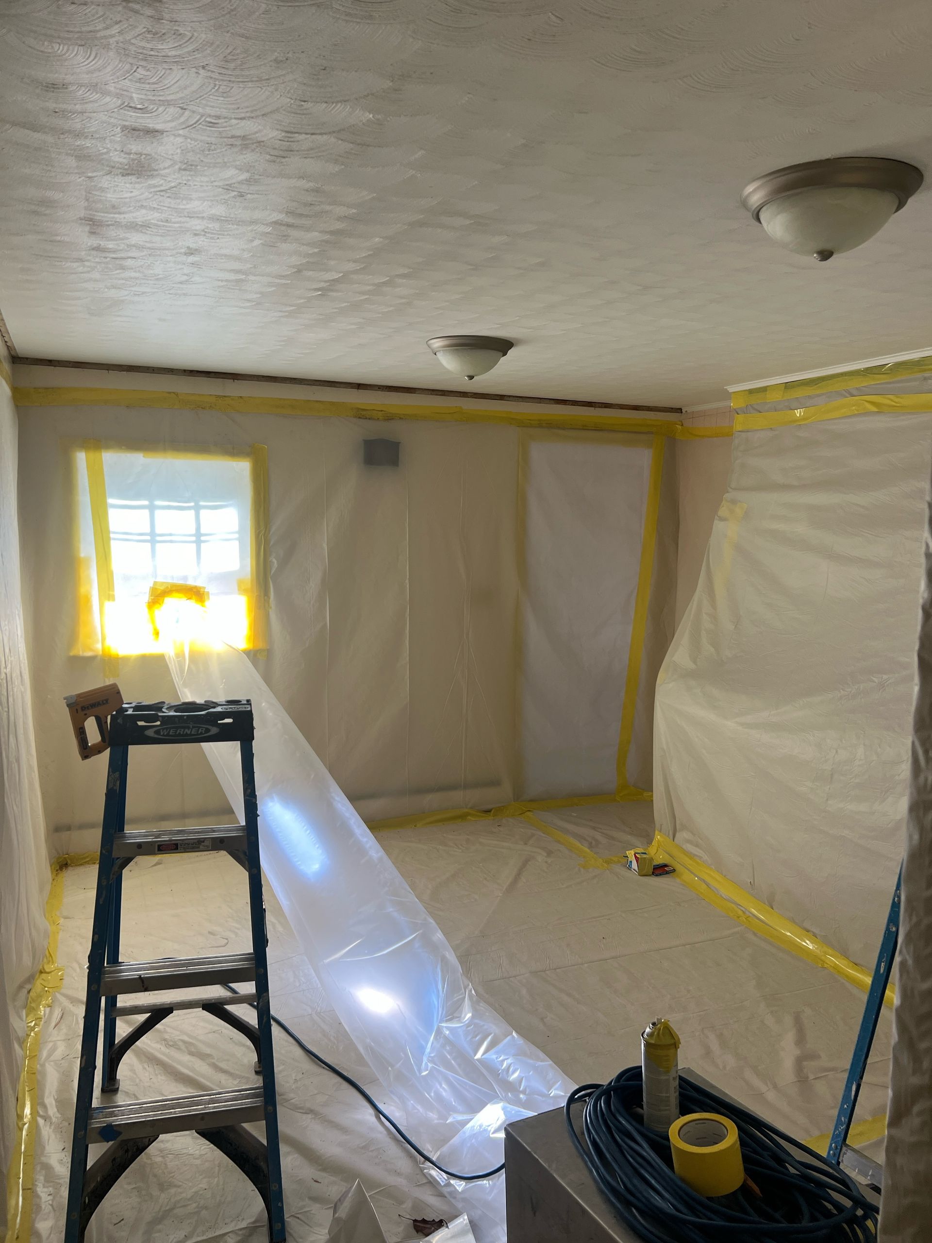 Room sealed with plastic sheeting and yellow tape during remediation. Ladder, lights, and a window are visible.