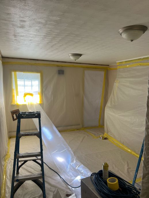 Room sealed with plastic sheeting, taped yellow, for mold remediation. A ladder stands near a window.