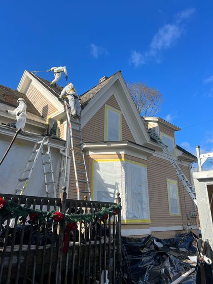 Workers painting a light-colored house with ladders, covering windows. Clear blue sky.