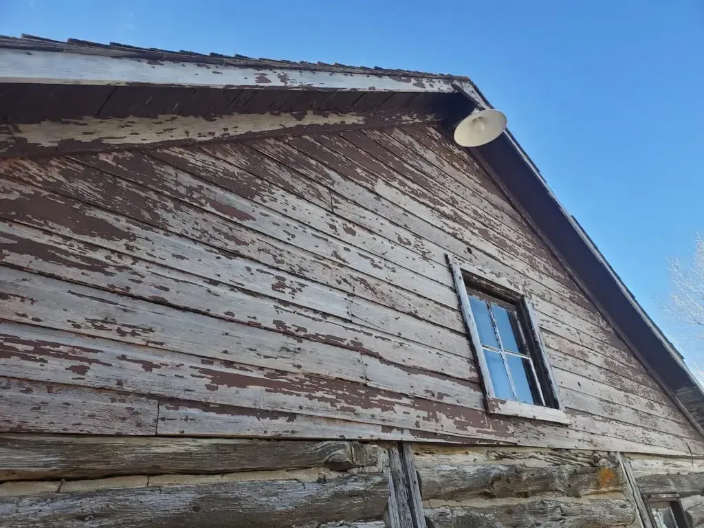 Weathered, brown wooden barn exterior with peeling paint, small window, and a satellite dish against a clear blue sky.