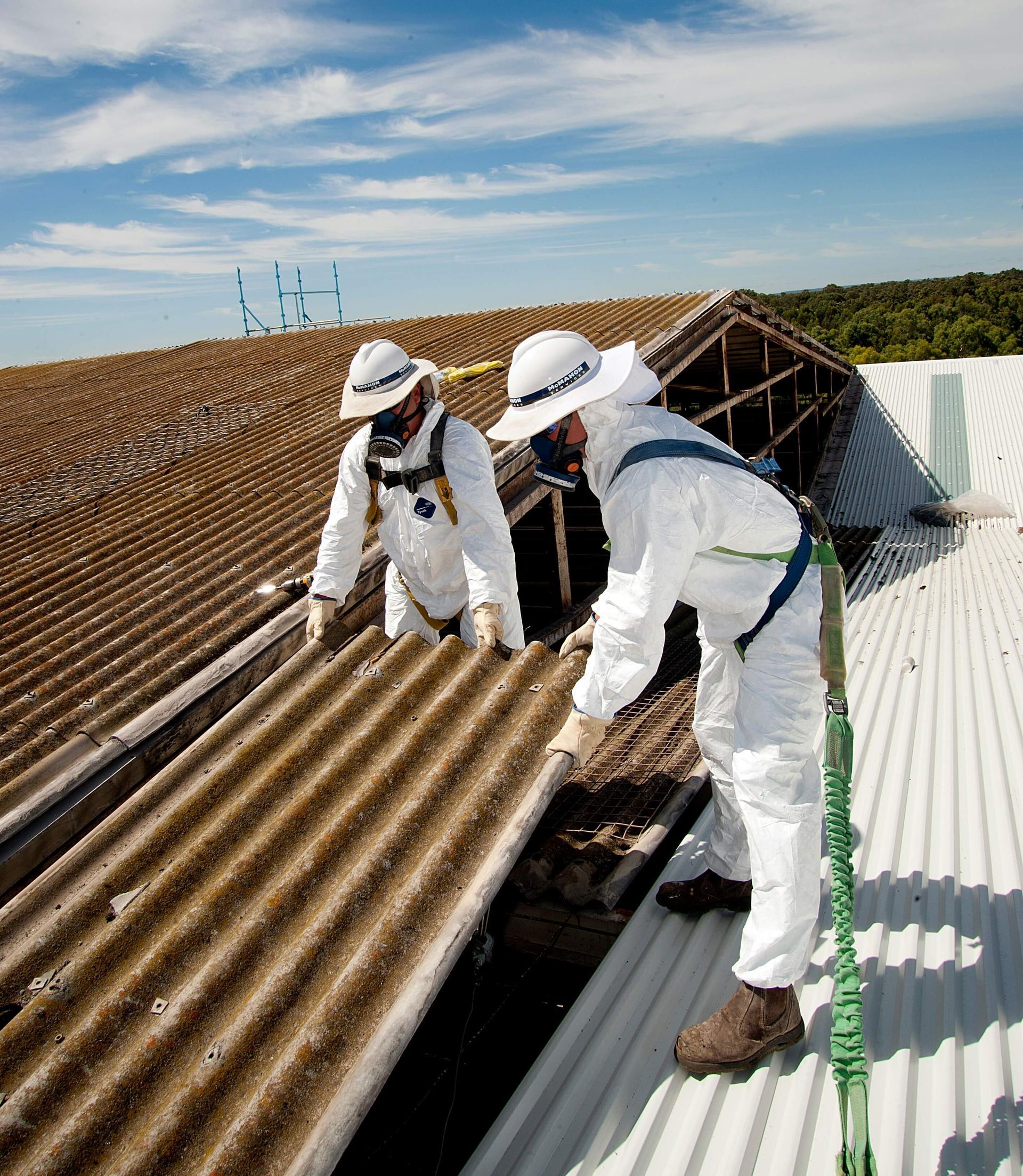 Two workers in protective suits remove asbestos roofing on a sunny day.