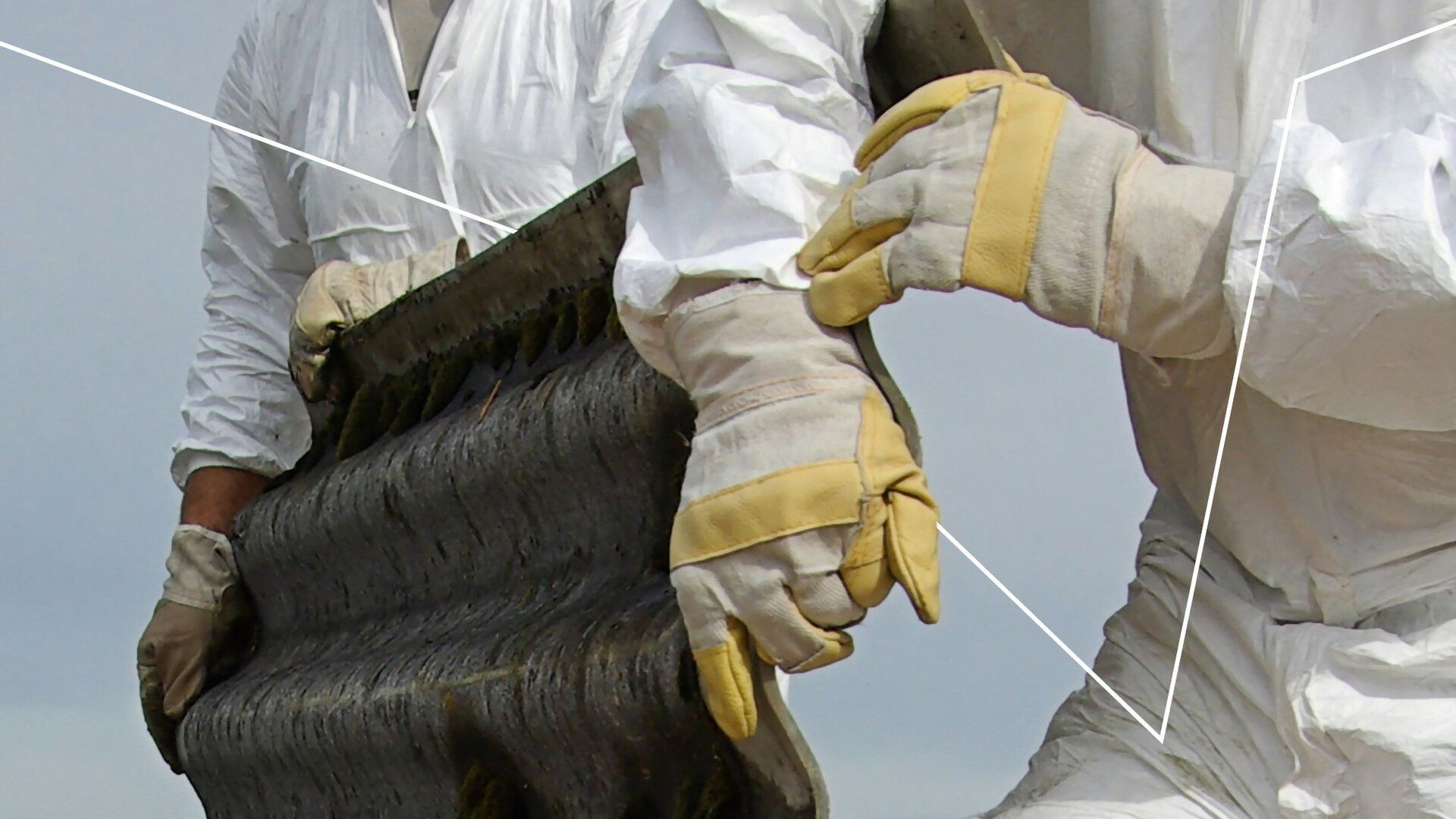 Workers in white suits and gloves holding a large, dark, possibly contaminated, sheet outdoors.