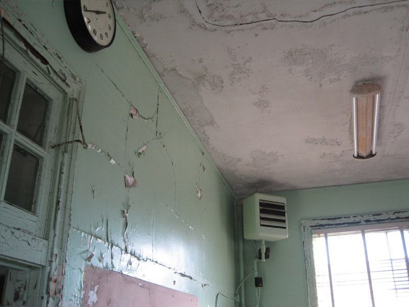 Cracked green walls and ceiling with peeling paint, a clock, and a window in a dilapidated room.