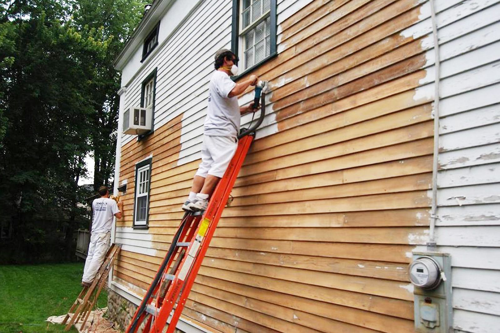 Two painters on ladders repaint a house's wooden siding. Brown wood contrasts with white siding.