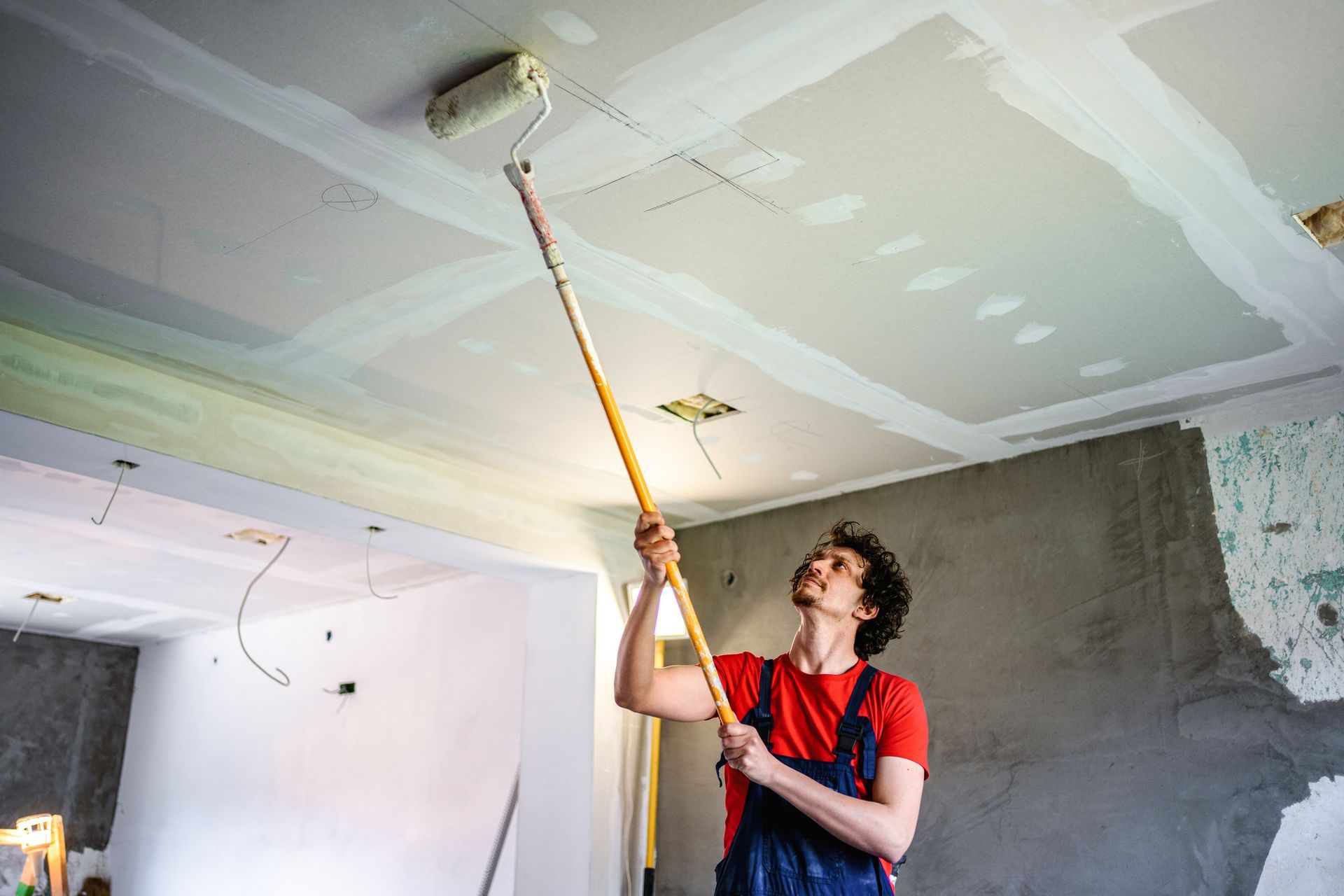 A construction worker is painting an apartment ceiling with a paint roller.