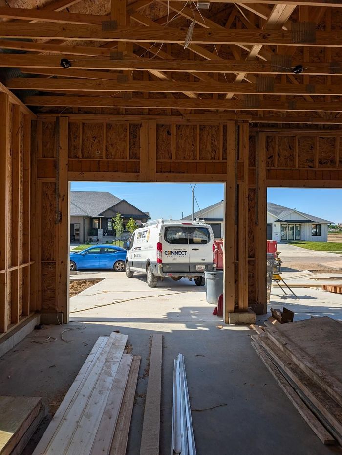 Interior of a framed garage under construction, looking out to a driveway with a white van and neighboring houses.