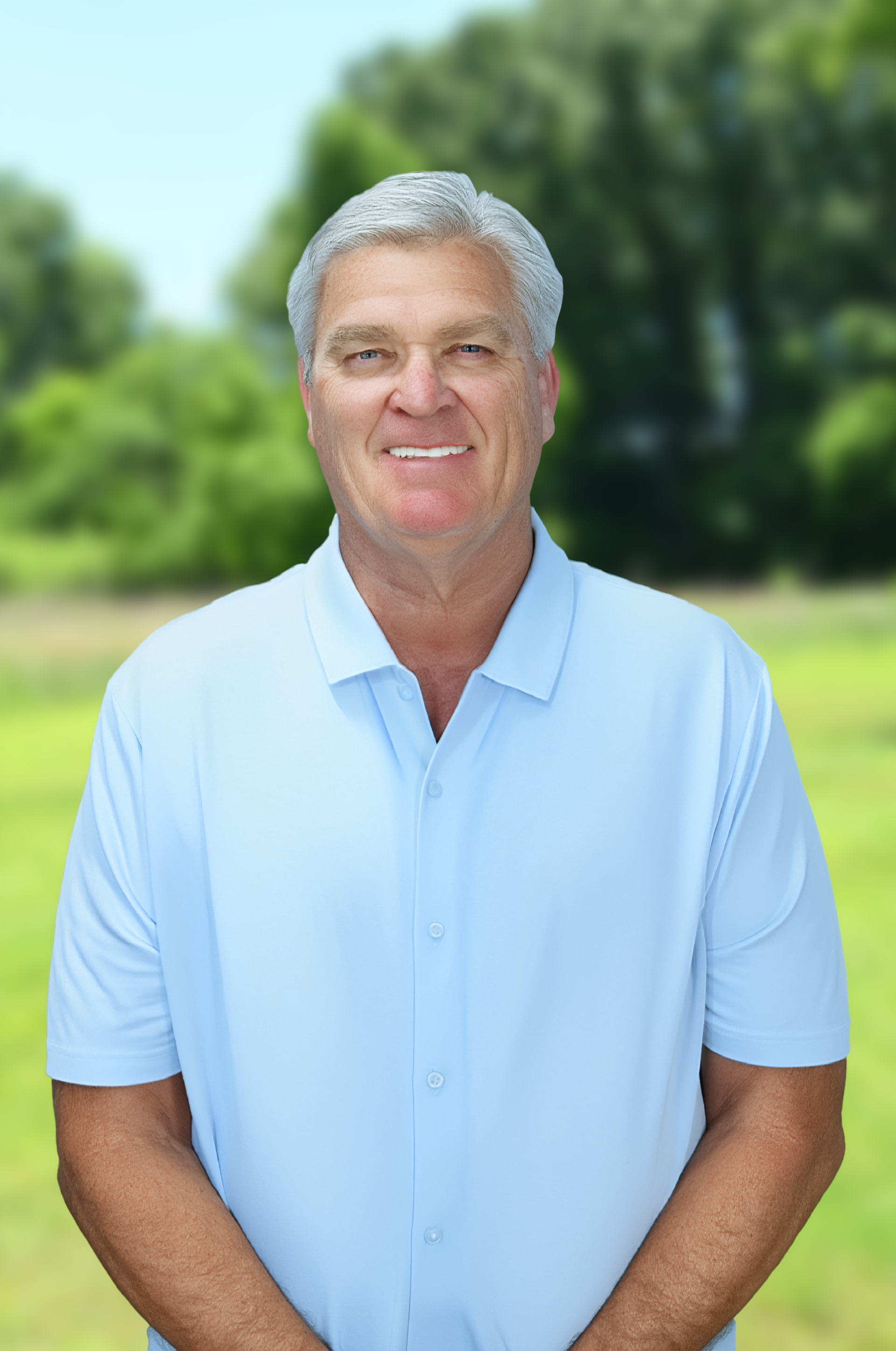Smiling man in a light blue polo standing by a pond with a fountain and building in the background