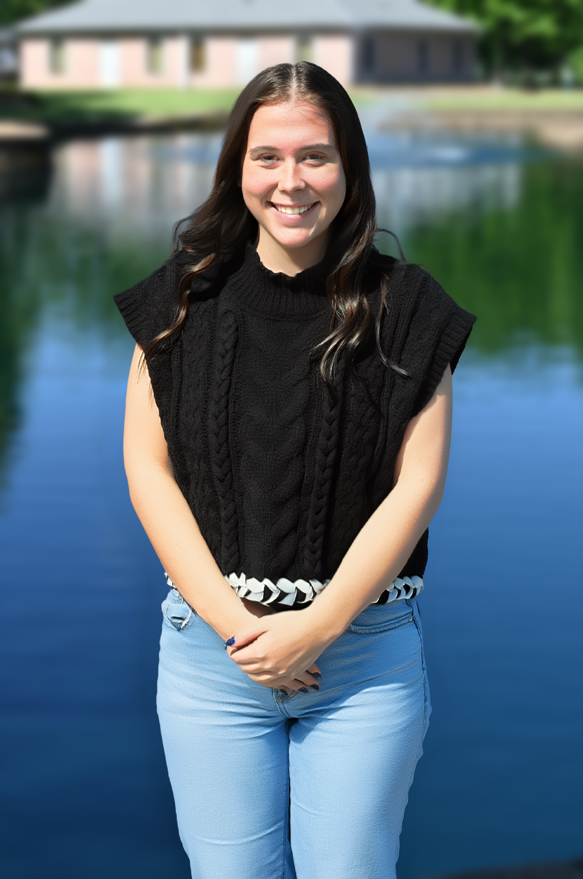 Young woman in a black top and jeans standing by a pond with a building in the background