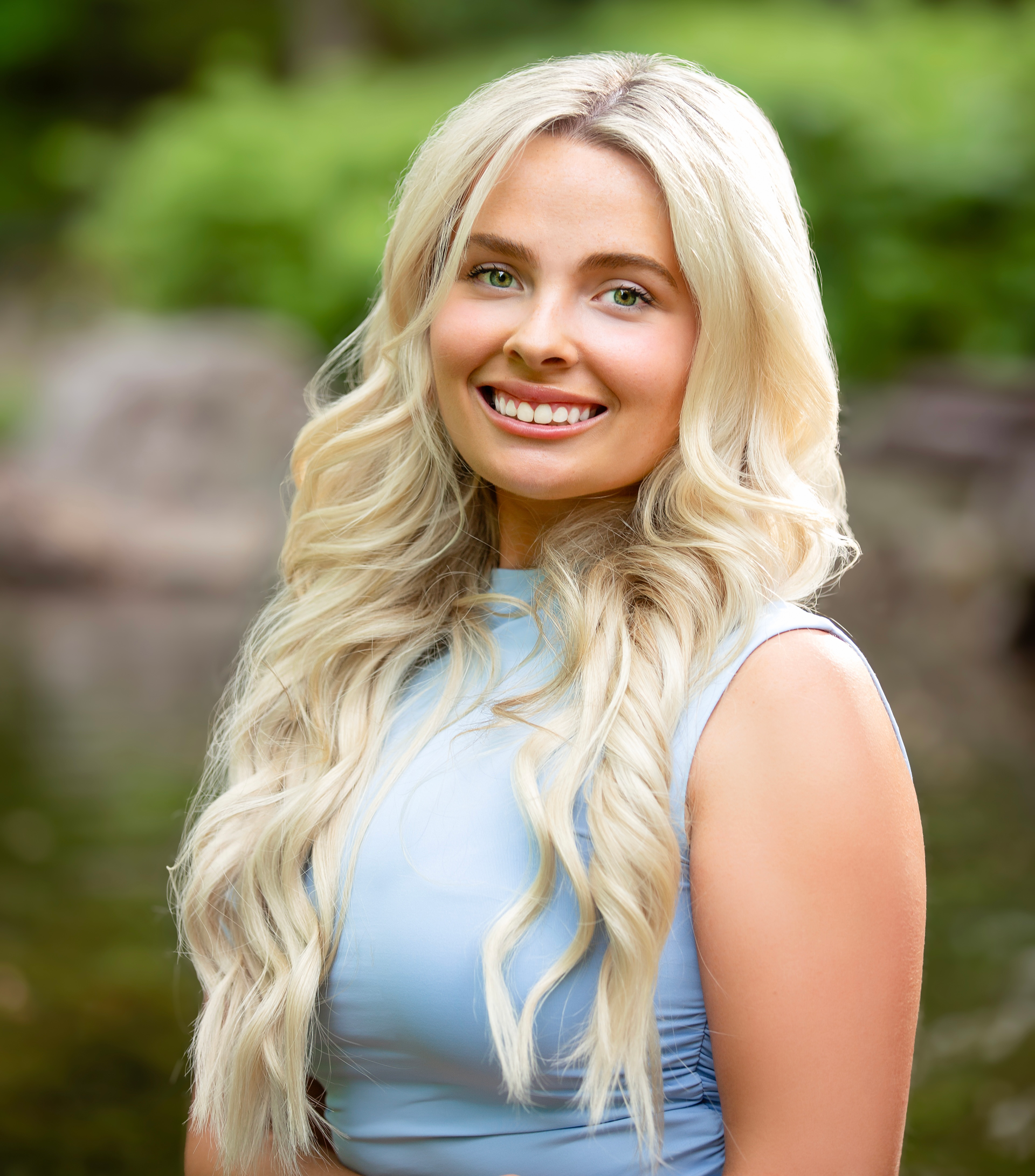 Smiling blonde woman in a light blue sleeveless dress standing outdoors with a blurred green background