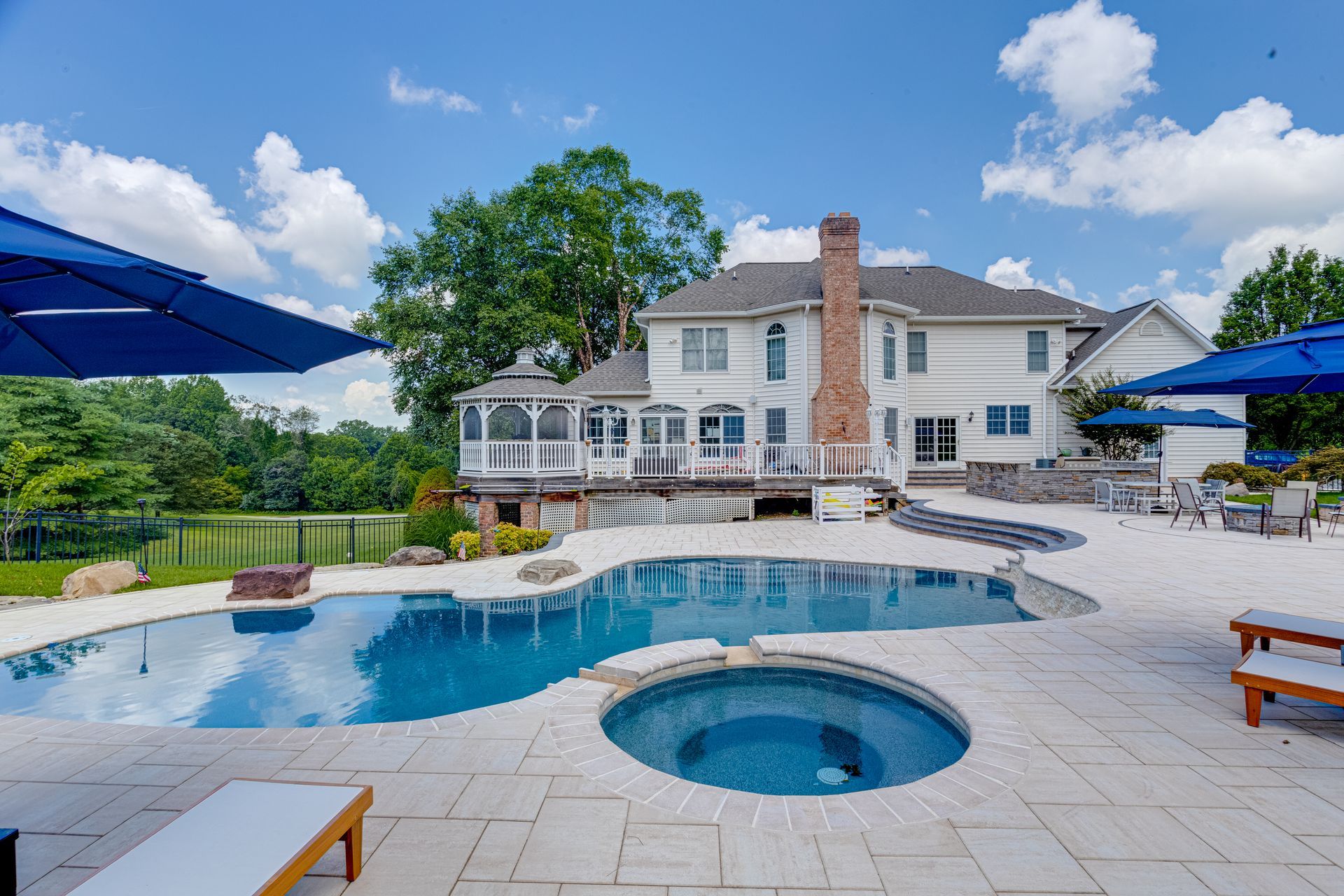 Backyard pool and hot tub in front of a large white house with patio seating and blue umbrellas