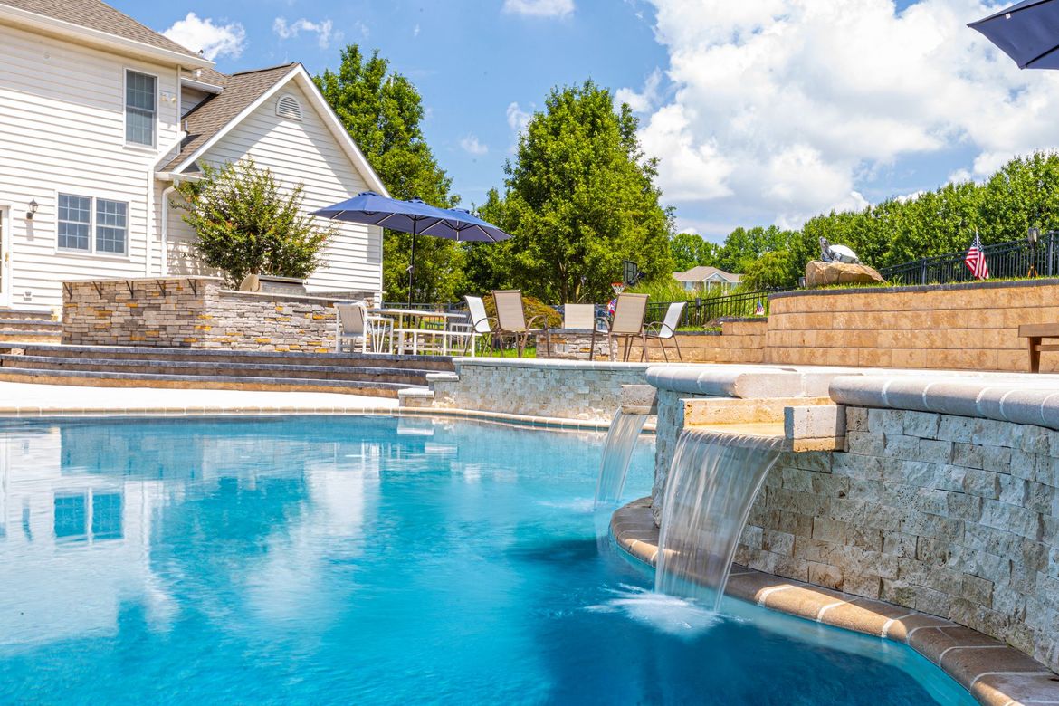 Pool area with waterfall, blue water, beige stone walls, and a white house under a blue sky.