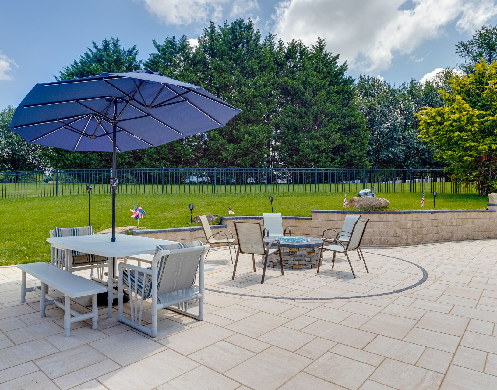 A patio with a dining table under a blue umbrella, a fire pit, and chairs on a stone-paved yard against a treeline.