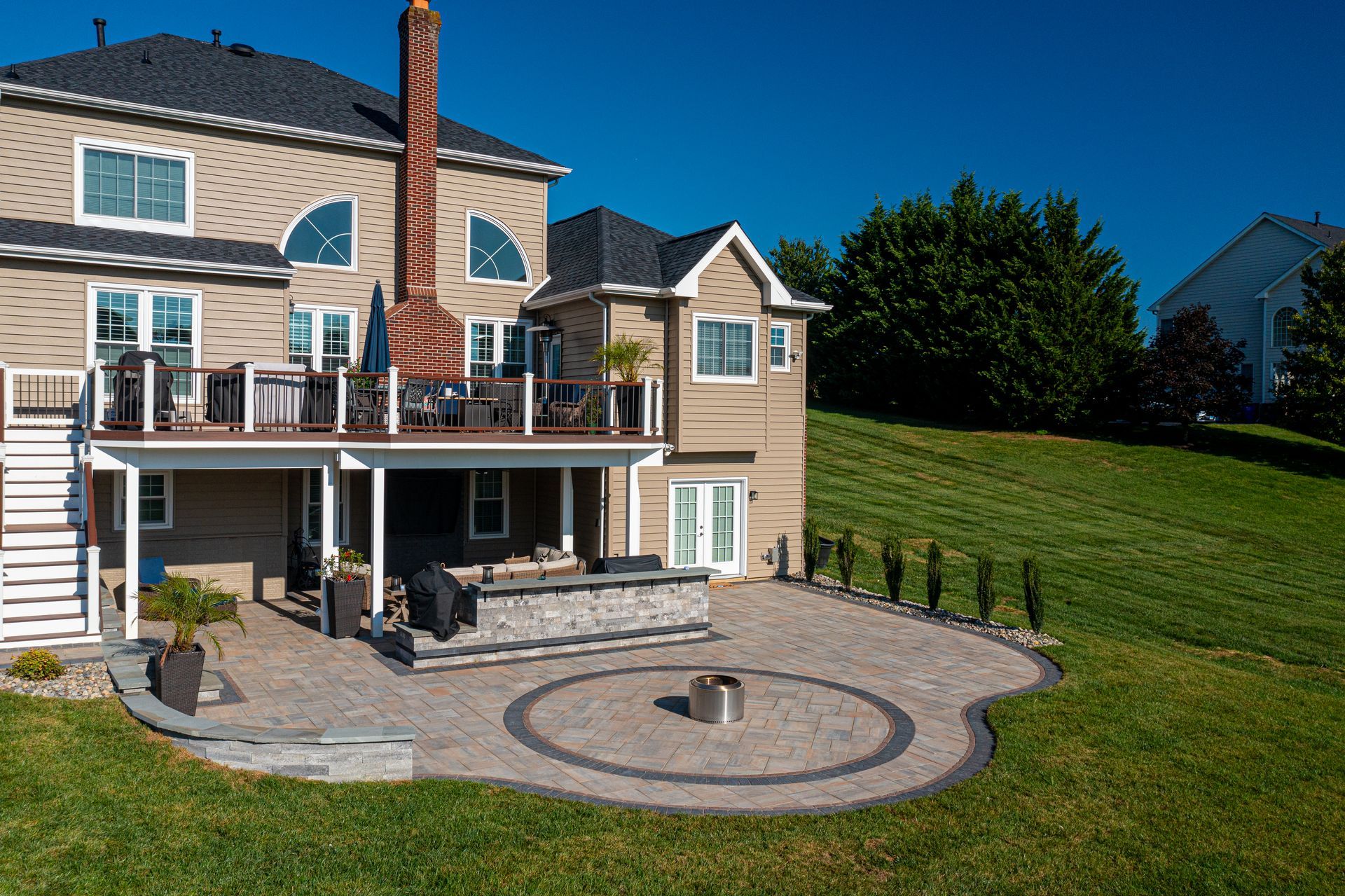 Backyard patio with fire pit, stone seating, and deck attached to a two-story beige house on a sunny day.