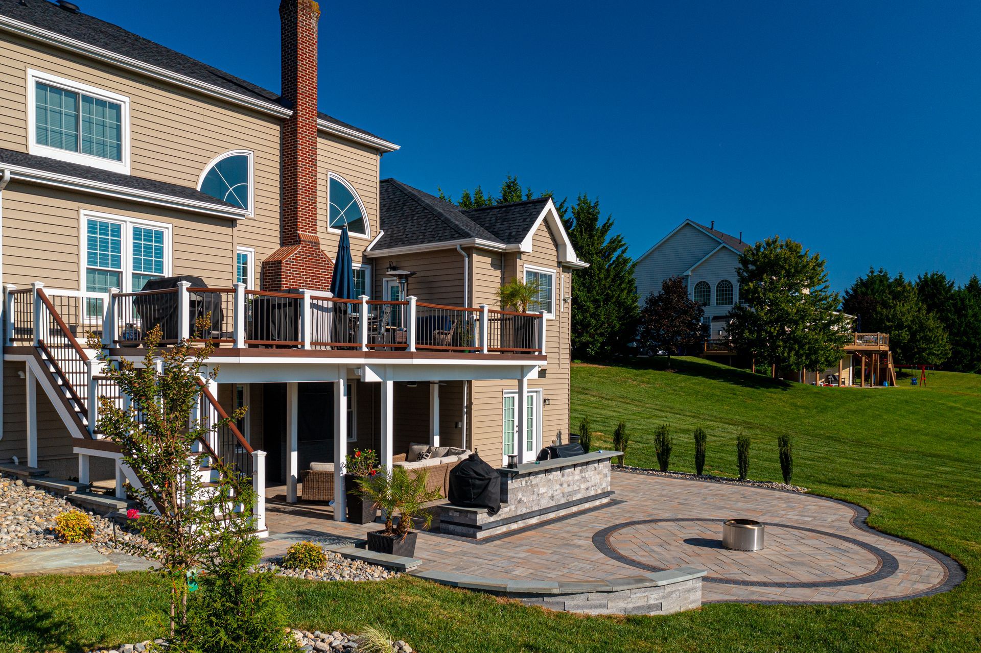 Backyard with a two-story house, a deck, a patio with a fire pit, and a grassy lawn on a sunny day.