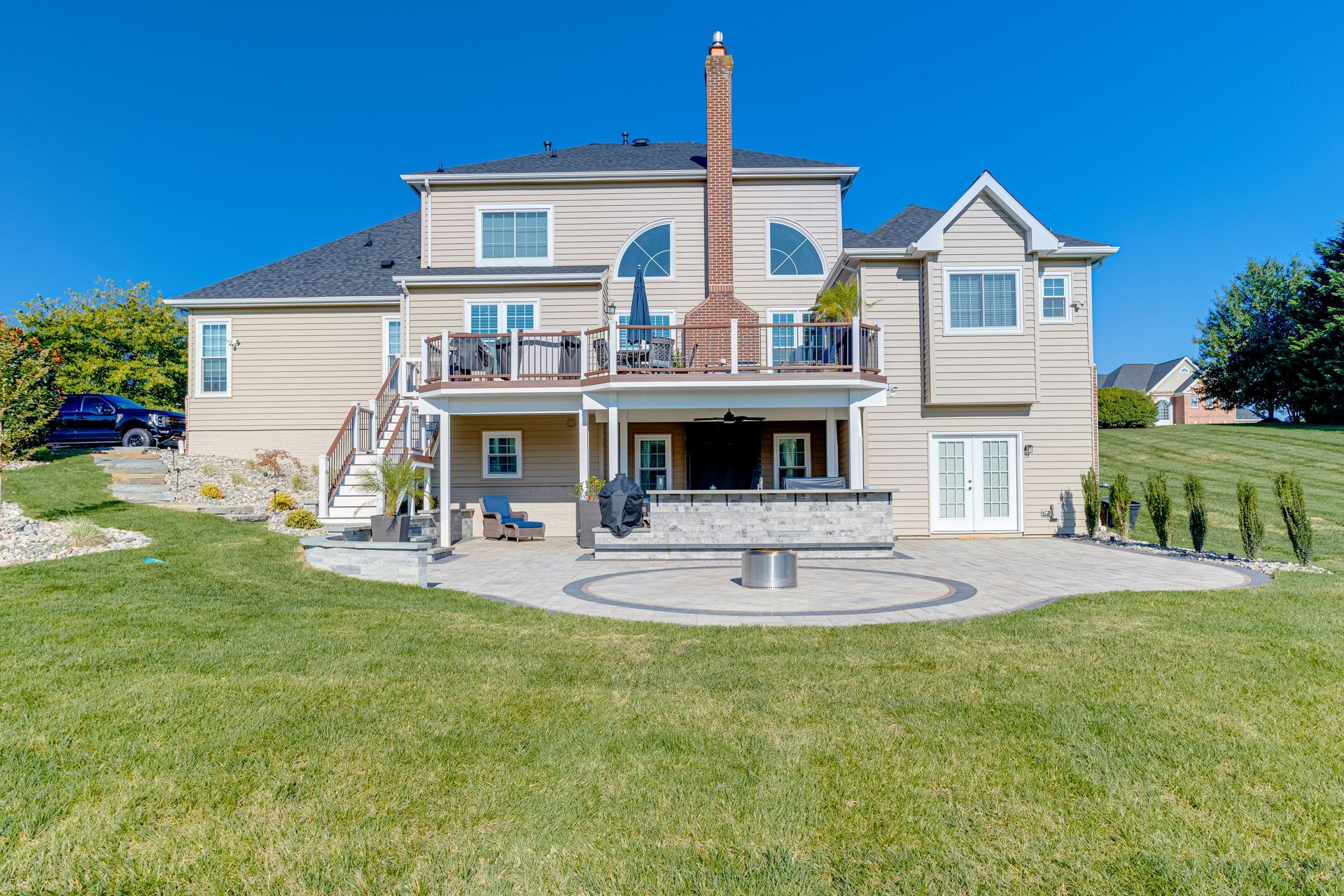 Beige two-story house with decks, patio, and chimney. Sunny day, clear blue sky, green lawn.