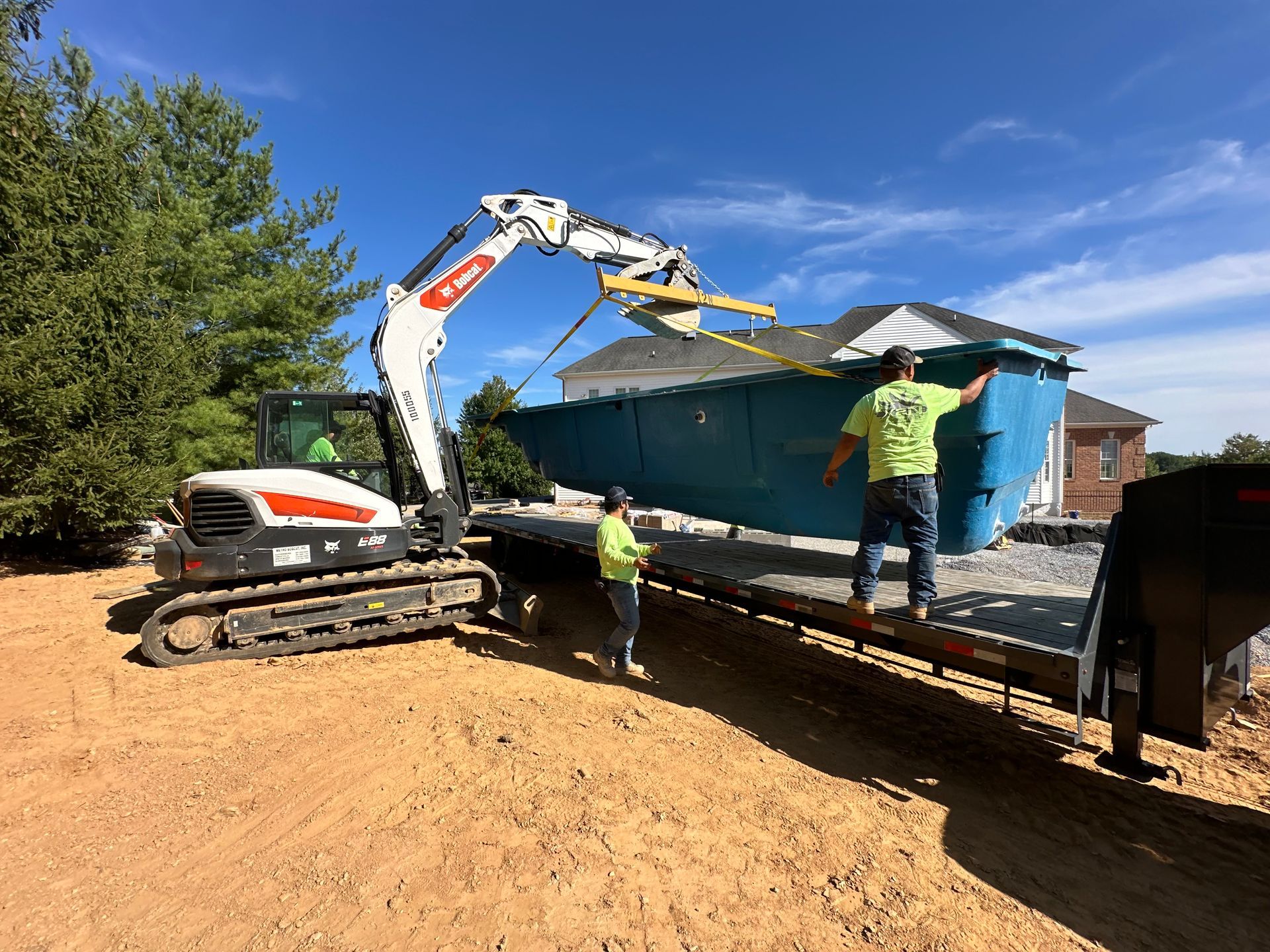 A man in a green shirt is standing next to a bulldozer carrying a pool on a trailer.