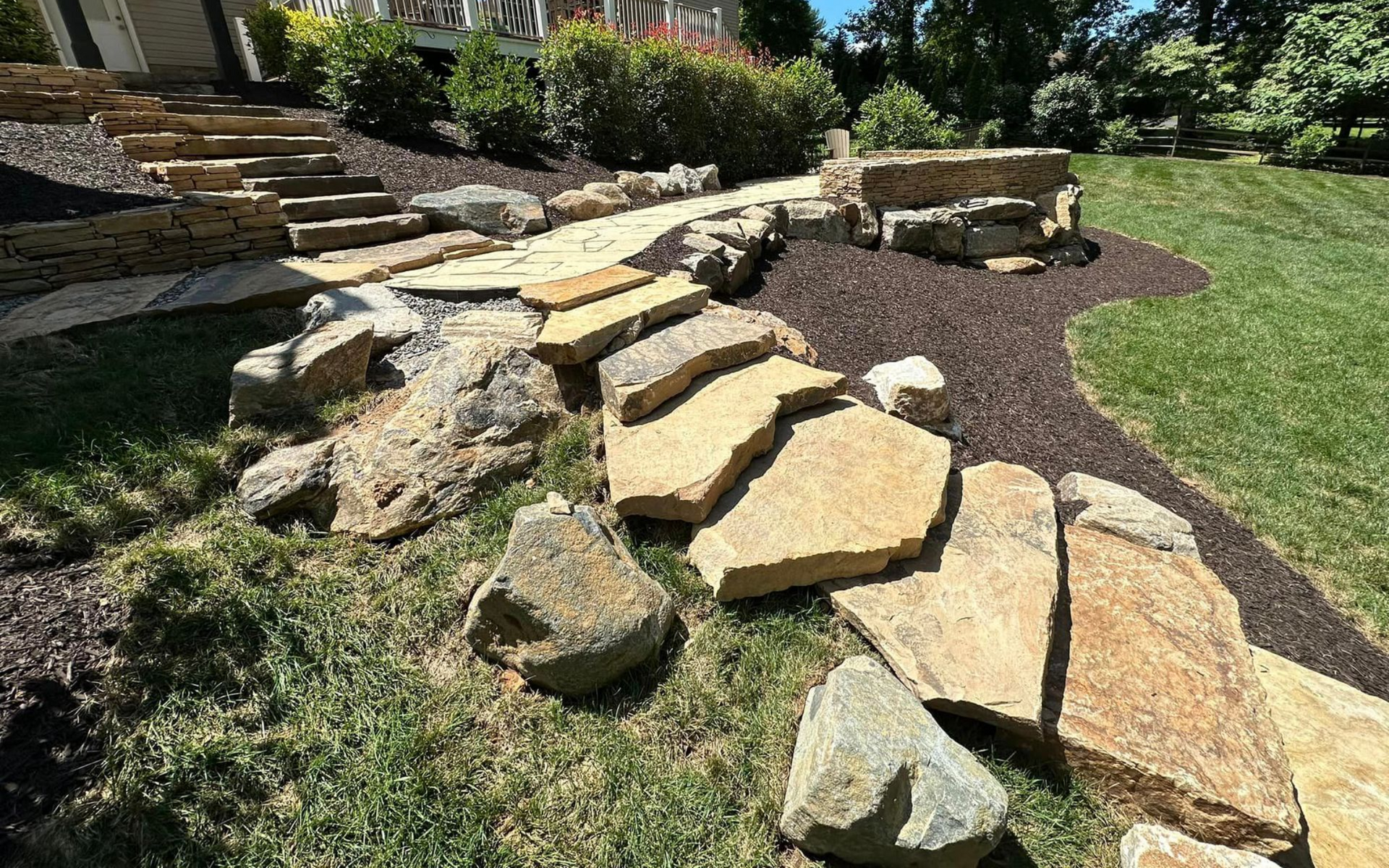 Stone pathway and steps ascending a sloped yard, bordered by rocks and mulch, with green grass.