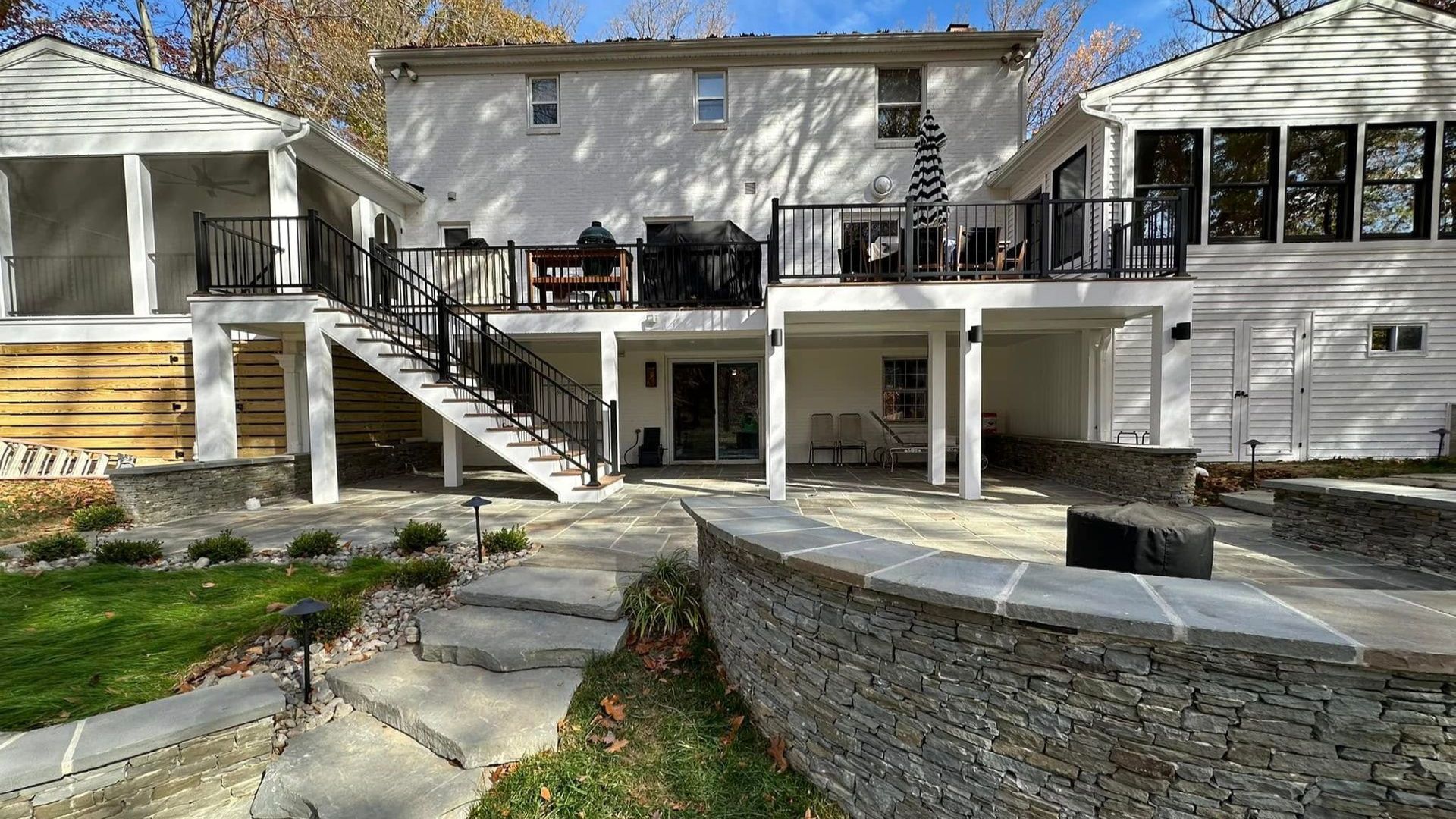 Backyard view of a white two-story house with decks, stairs, and a stone wall patio designed by X2N
