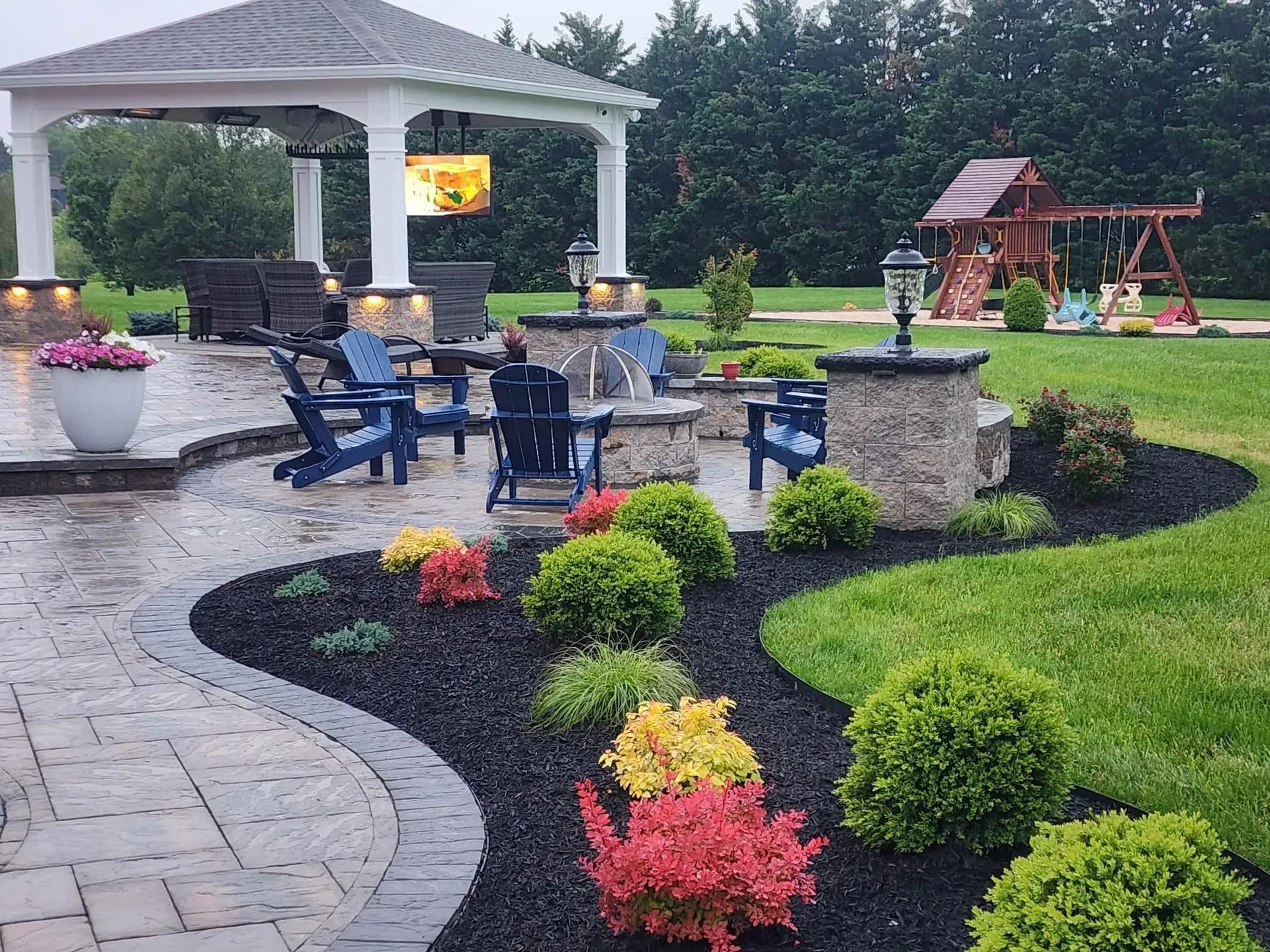 Backyard patio with gazebo, blue chairs, fire pit, and landscaped flower beds with black mulch.