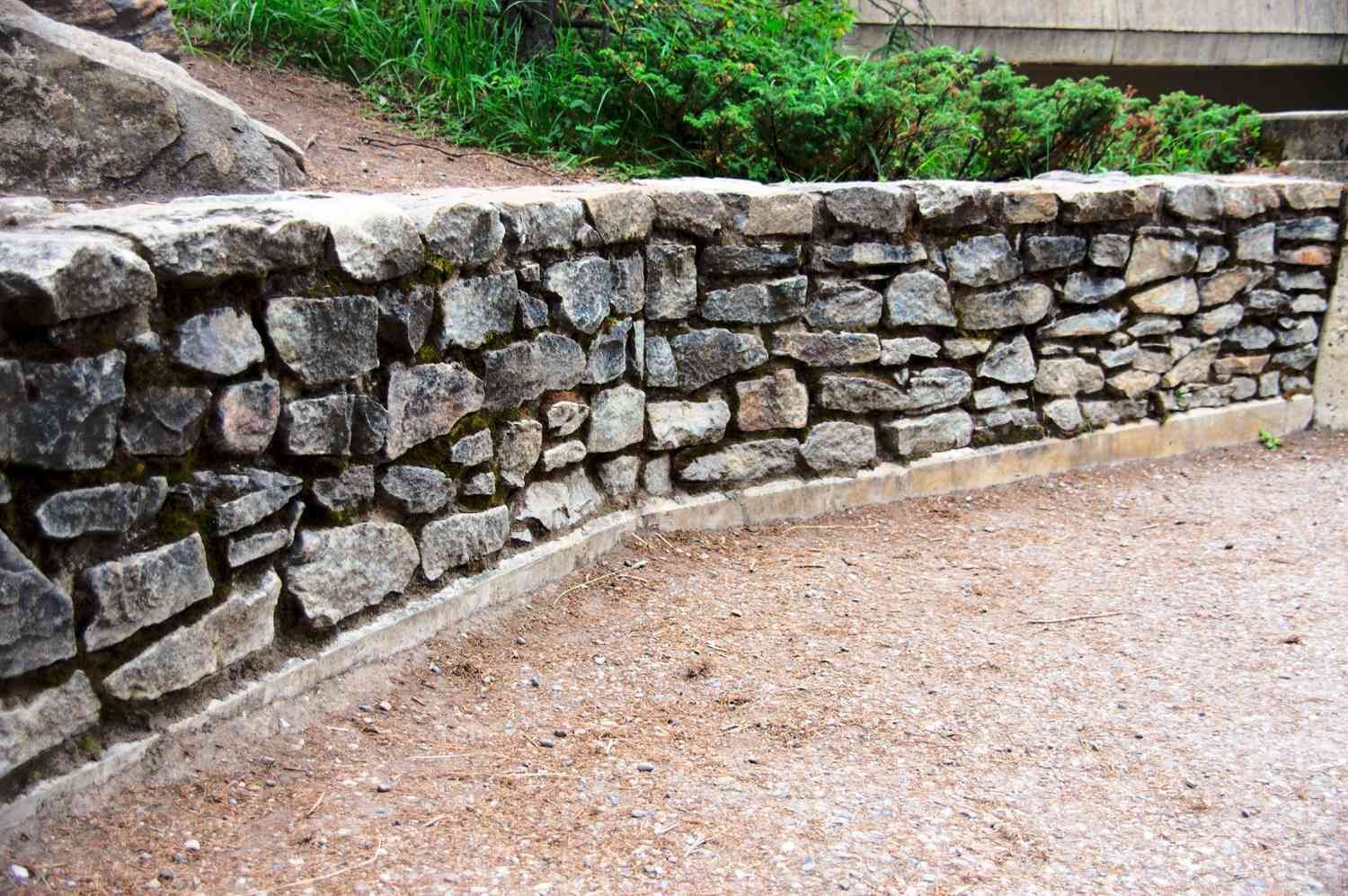Stone retaining wall with curved shape, next to gravel ground, greenery in background.