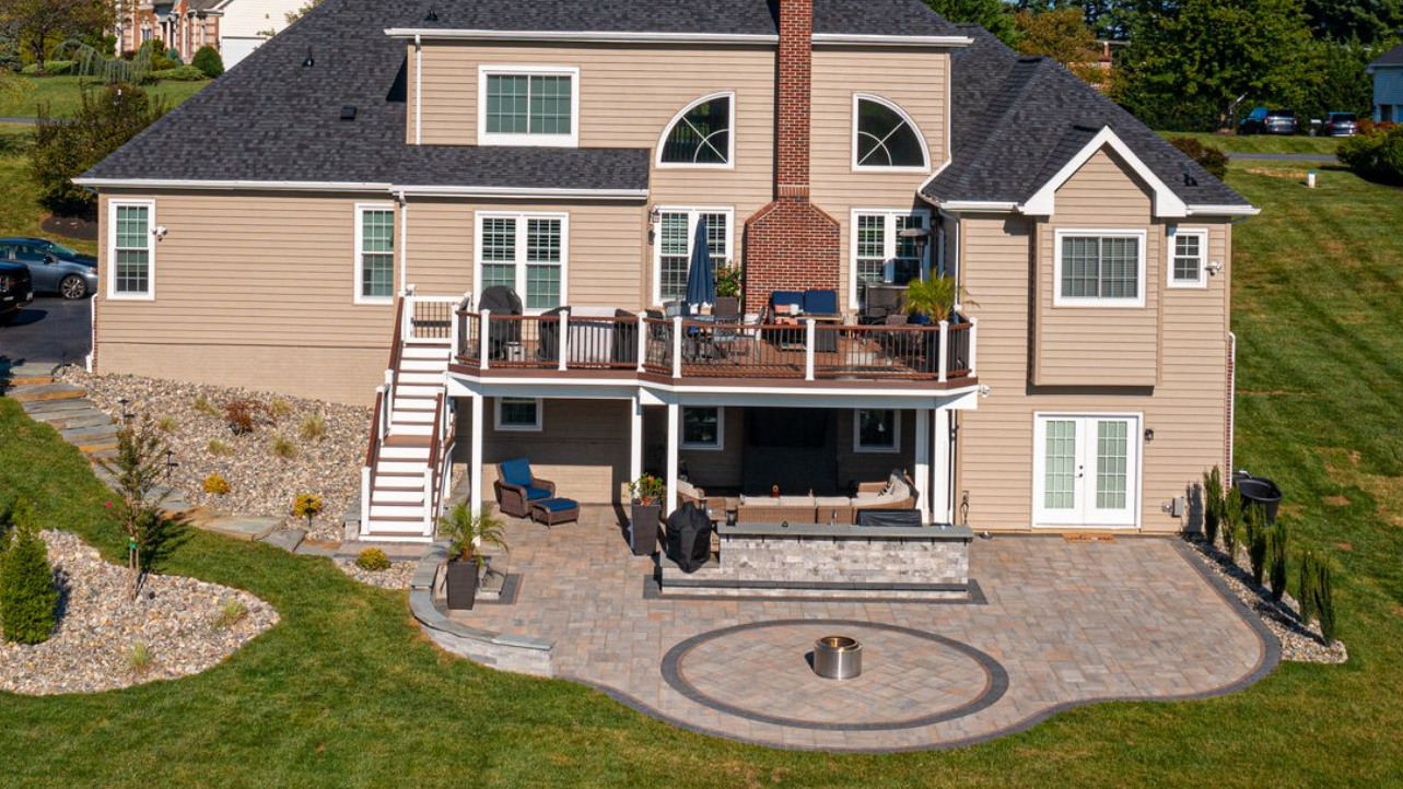 Suburban beige house with a raised deck, stairs, stone patio, and circular fire pit in the backyard