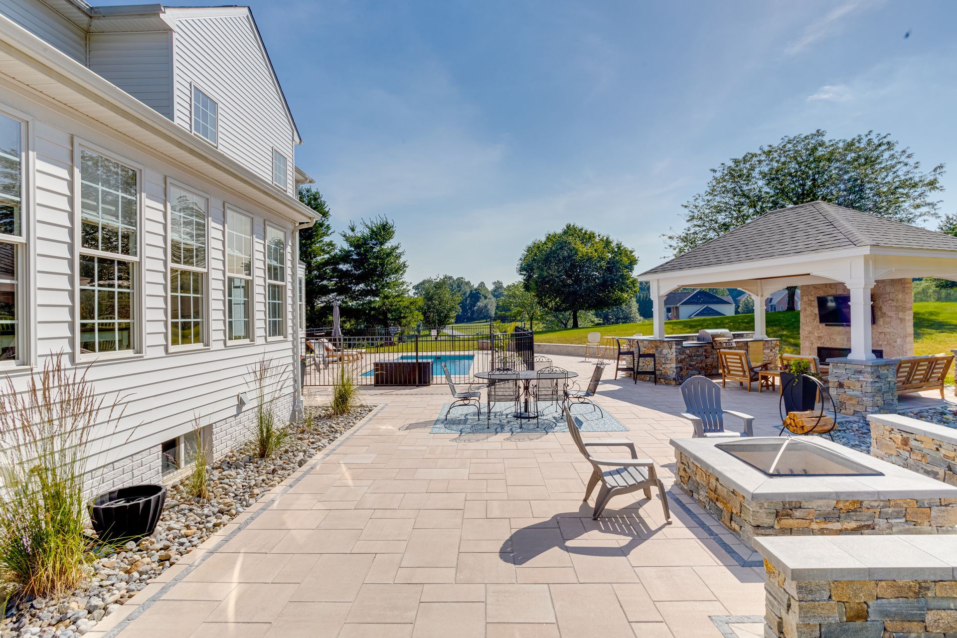 Backyard patio with a house, gazebo, pool, fire pit, and seating under a blue sky.