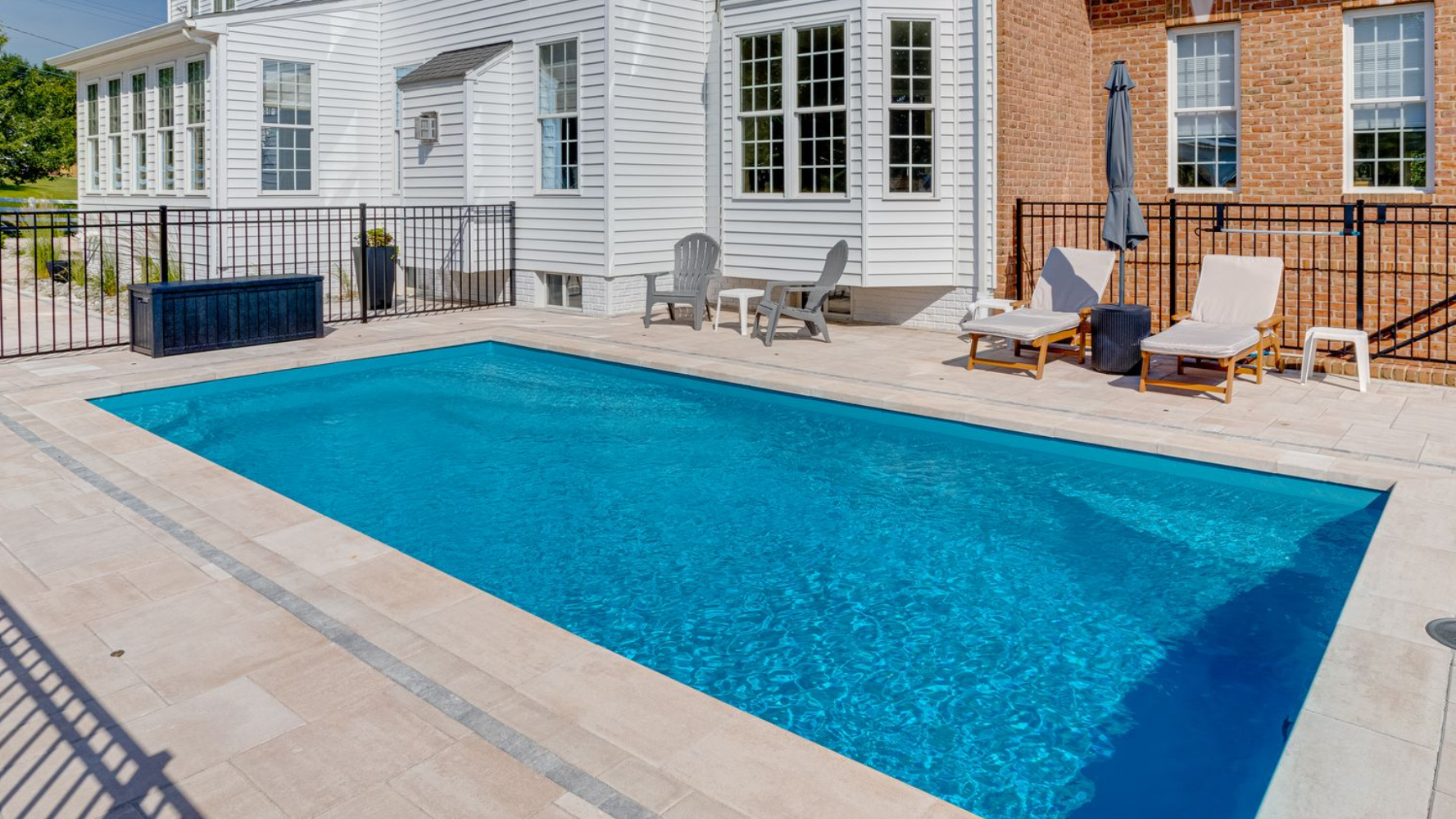 Rectangular blue swimming pool beside a white house with lounge chairs on a sunny patio