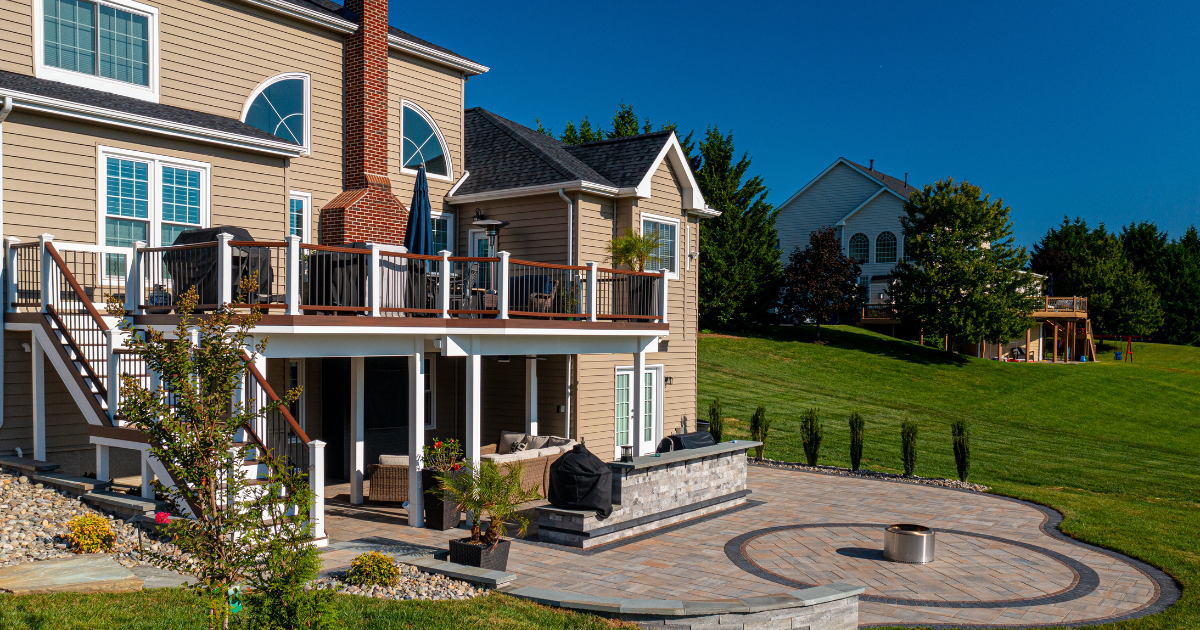 Brick house with a deck overlooking a landscaped backyard, stone patio, and green lawn under a blue sky