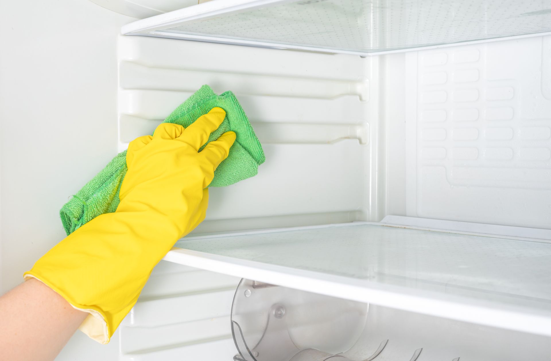 A person wearing yellow gloves is cleaning an empty refrigerator with a green cloth.